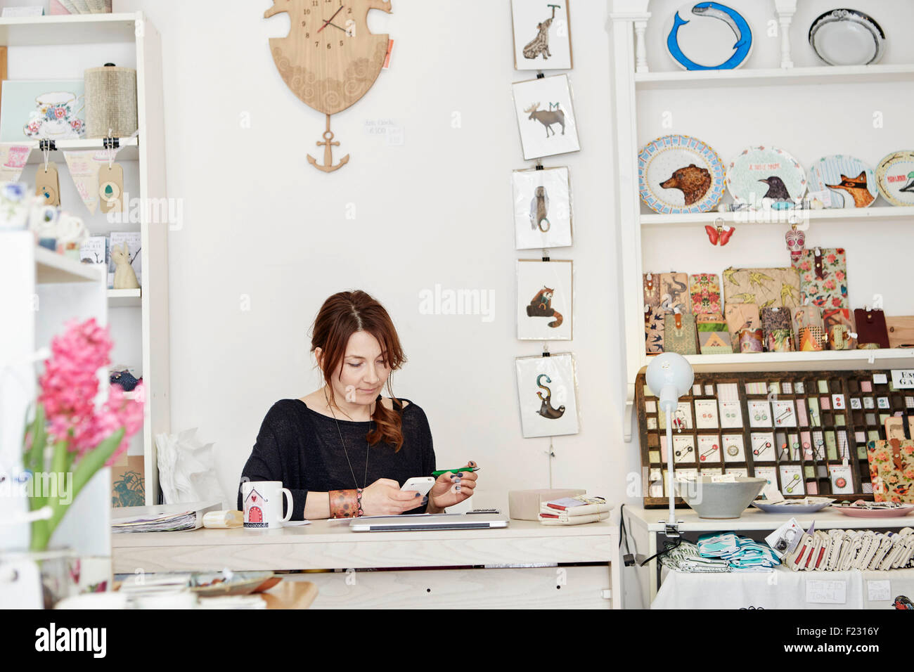 Eine Frau sitzen an einem Schreibtisch in einem kleinen Souvenir-Shop, den Papierkram zu tun, die Geschäftsführung, mit einem Laptop und einem Smartphone. Stockfoto