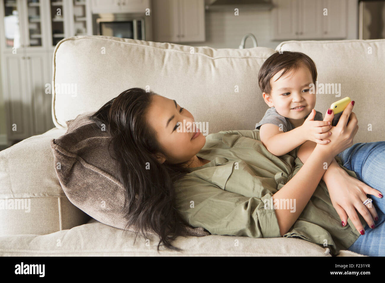 Frau liegend auf dem Sofa, lächelnd, kuscheln mit ihrem kleinen Sohn und mit Blick auf ein Handy. Stockfoto