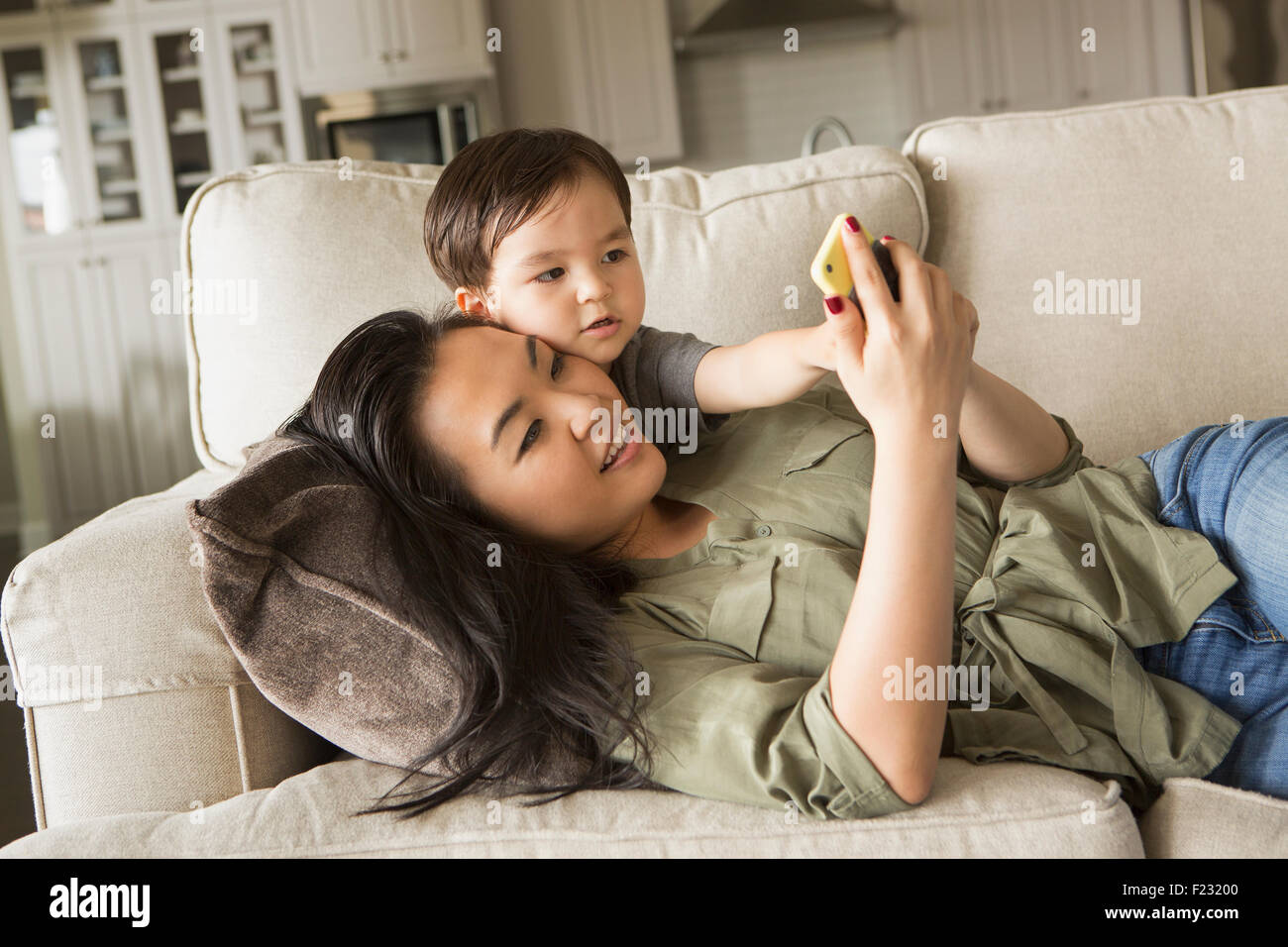 Frau liegend auf dem Sofa, lächelnd, kuscheln mit ihrem kleinen Sohn und mit Blick auf ein Handy
