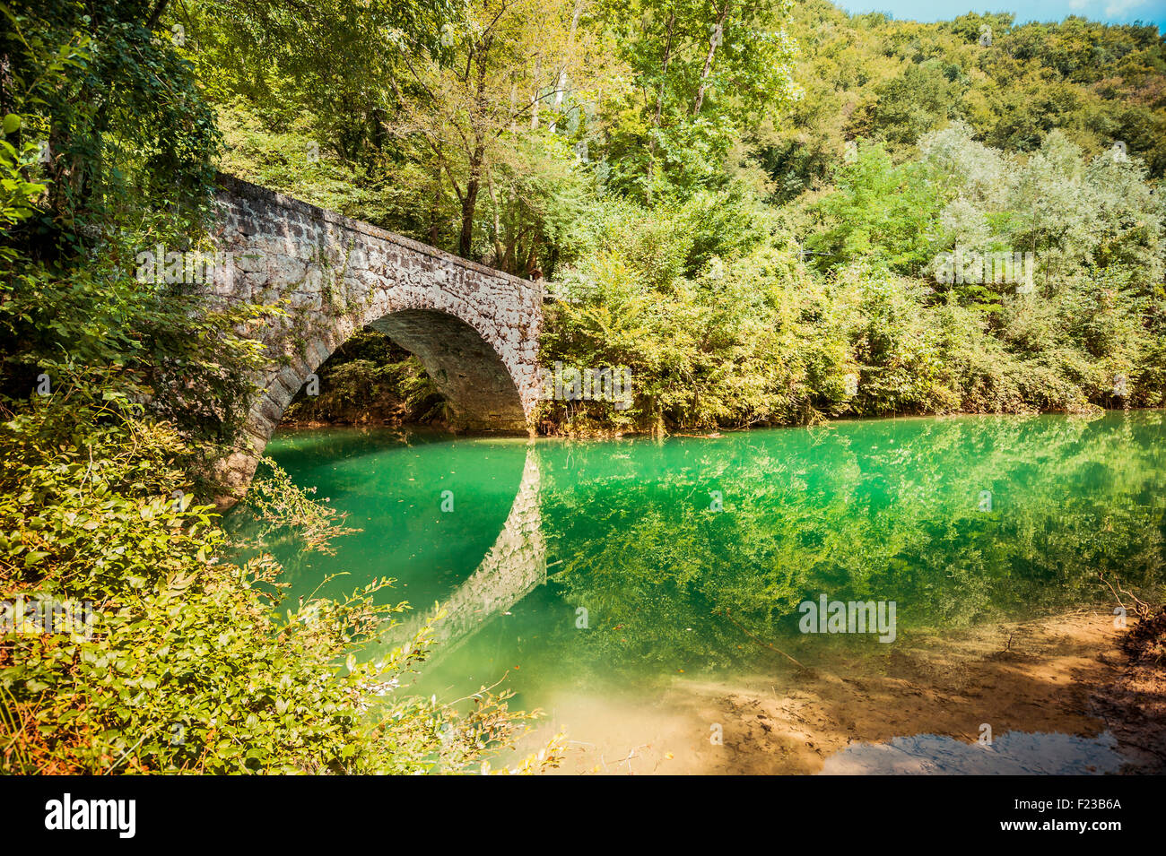 Alte Brücke von smaragdgrünen Wasser unter den Bäumen und ein Stück vom blauen Himmel Stockfoto