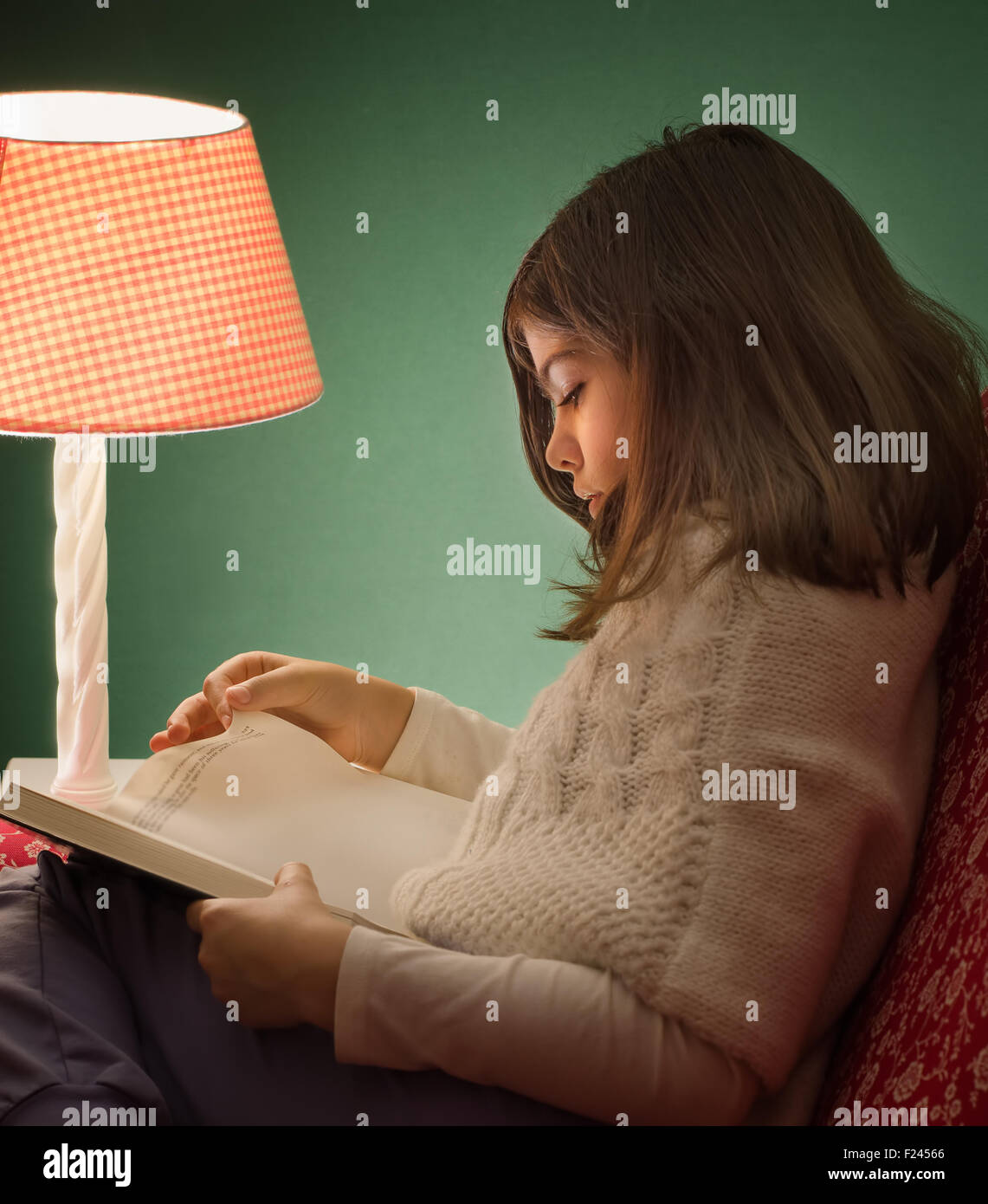 Kleines Mädchen, ein Buch lesen, unter rosa Lampe, mit grünem Hintergrund, in einer kalten Nacht mit Pullover. Stockfoto