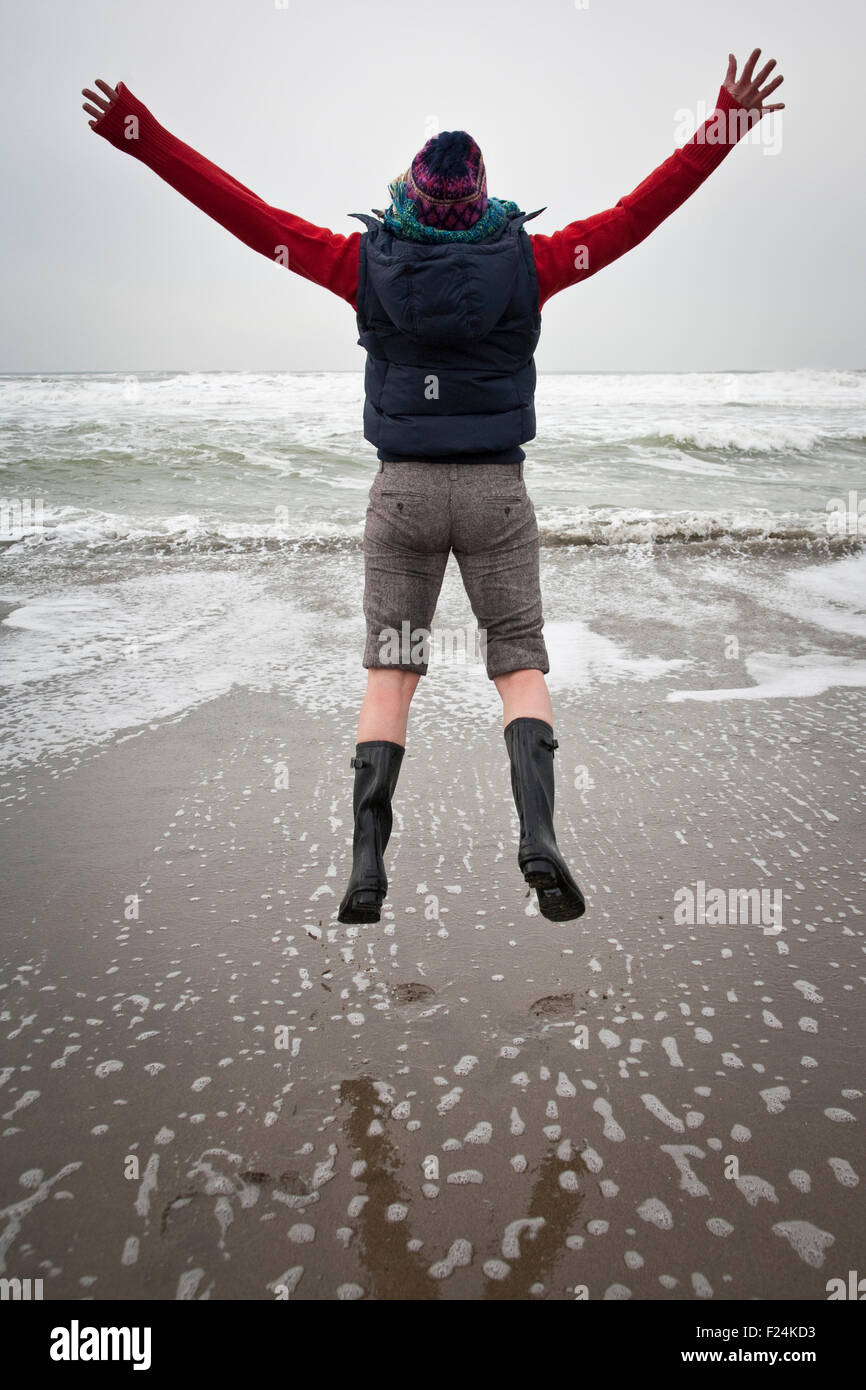 Junge Frau springt mit zurück in die Kamera und die Hände ausgestreckt auf einem Strand direkt am Meer an einem nebeligen Tag Stockfoto