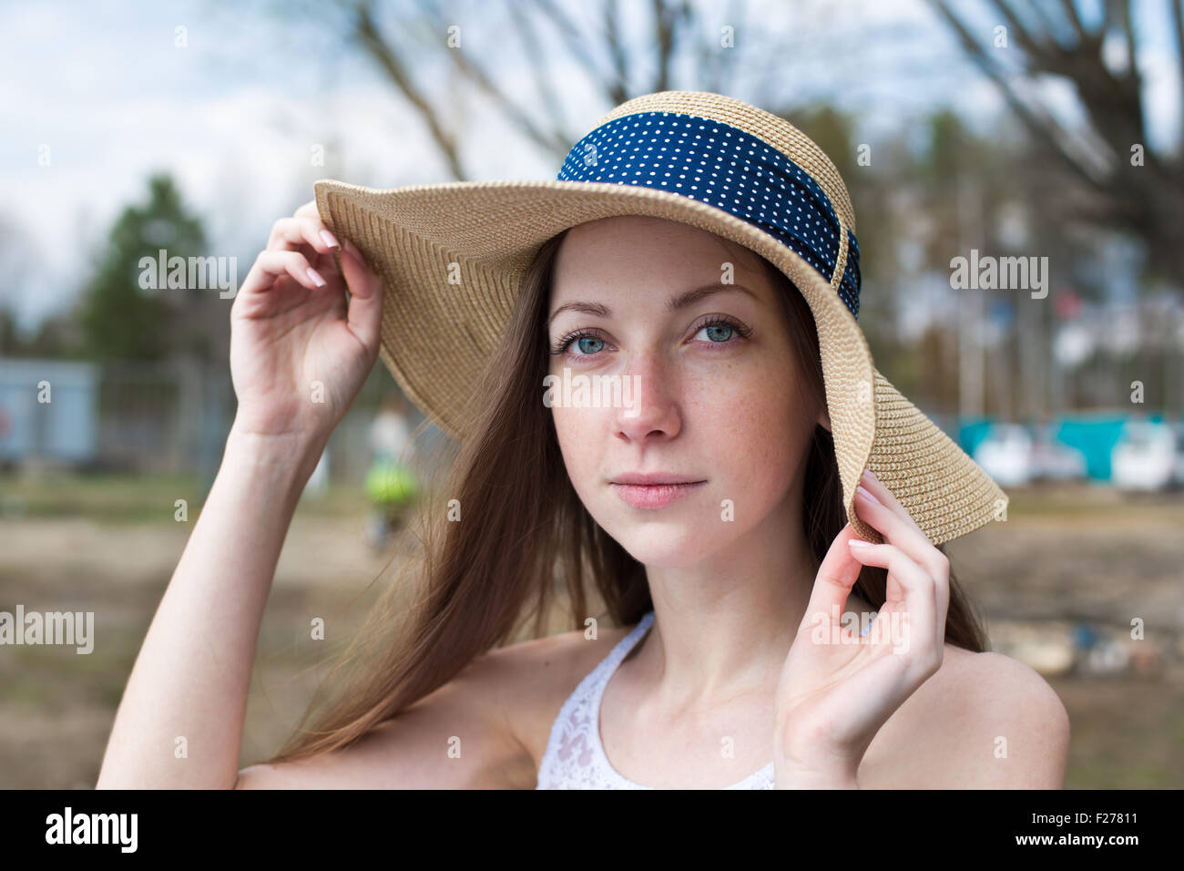 Sommersprossige glückliches Mädchen im weißen Kleid hält Stroh Hut und Blick in die Kamera bei windigem Wetter Stockfoto