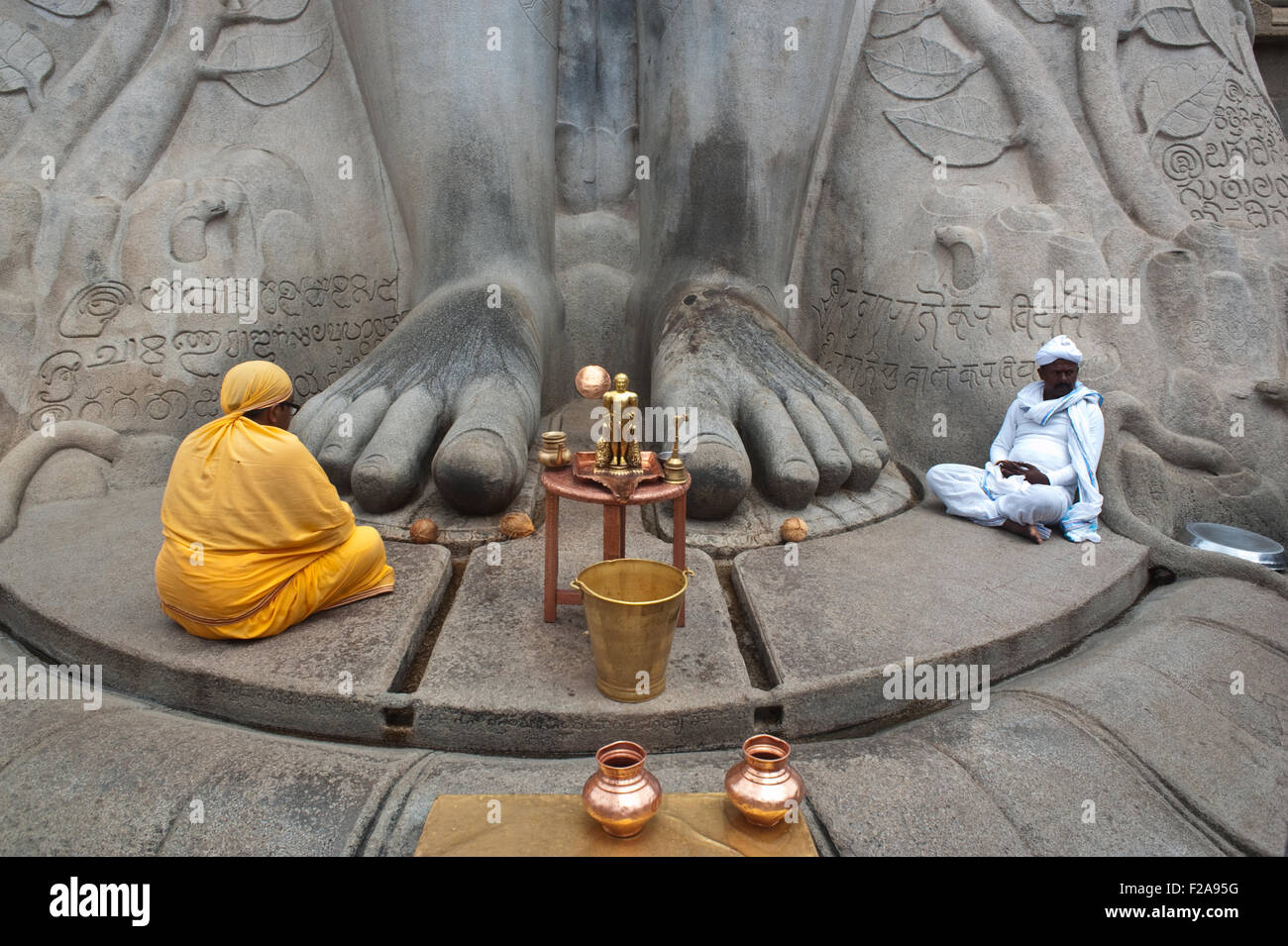 Jaïn Pilger (der Mann in Orange gekleidet) und ein Priester sitzen an den Füßen der Statue des Gomateshwara (Indien) Stockfoto
