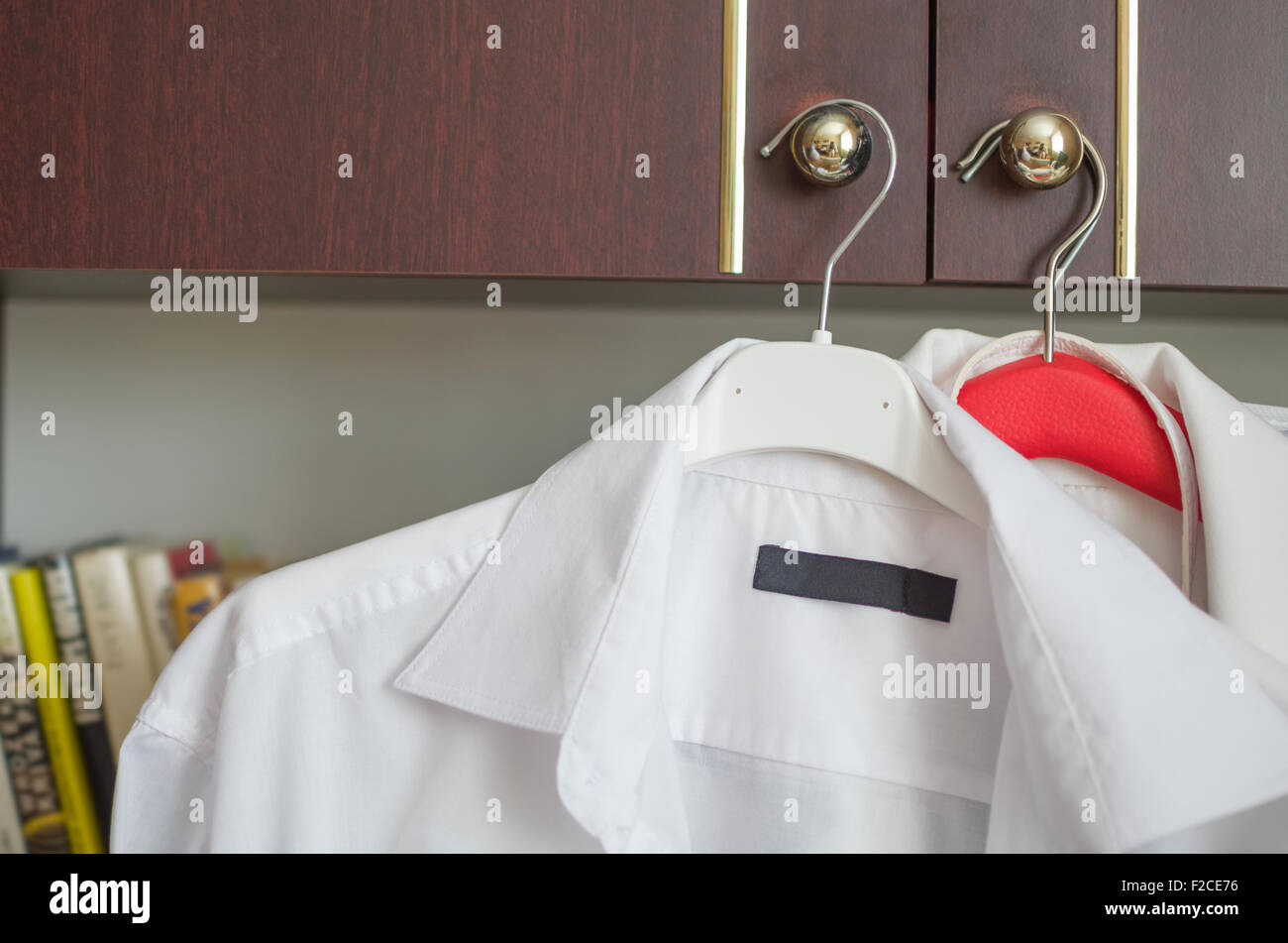 Weiße Hemden auf Kleiderbügeln auf Kabinetthandgriffe vor Bücherregal platziert Stockfoto