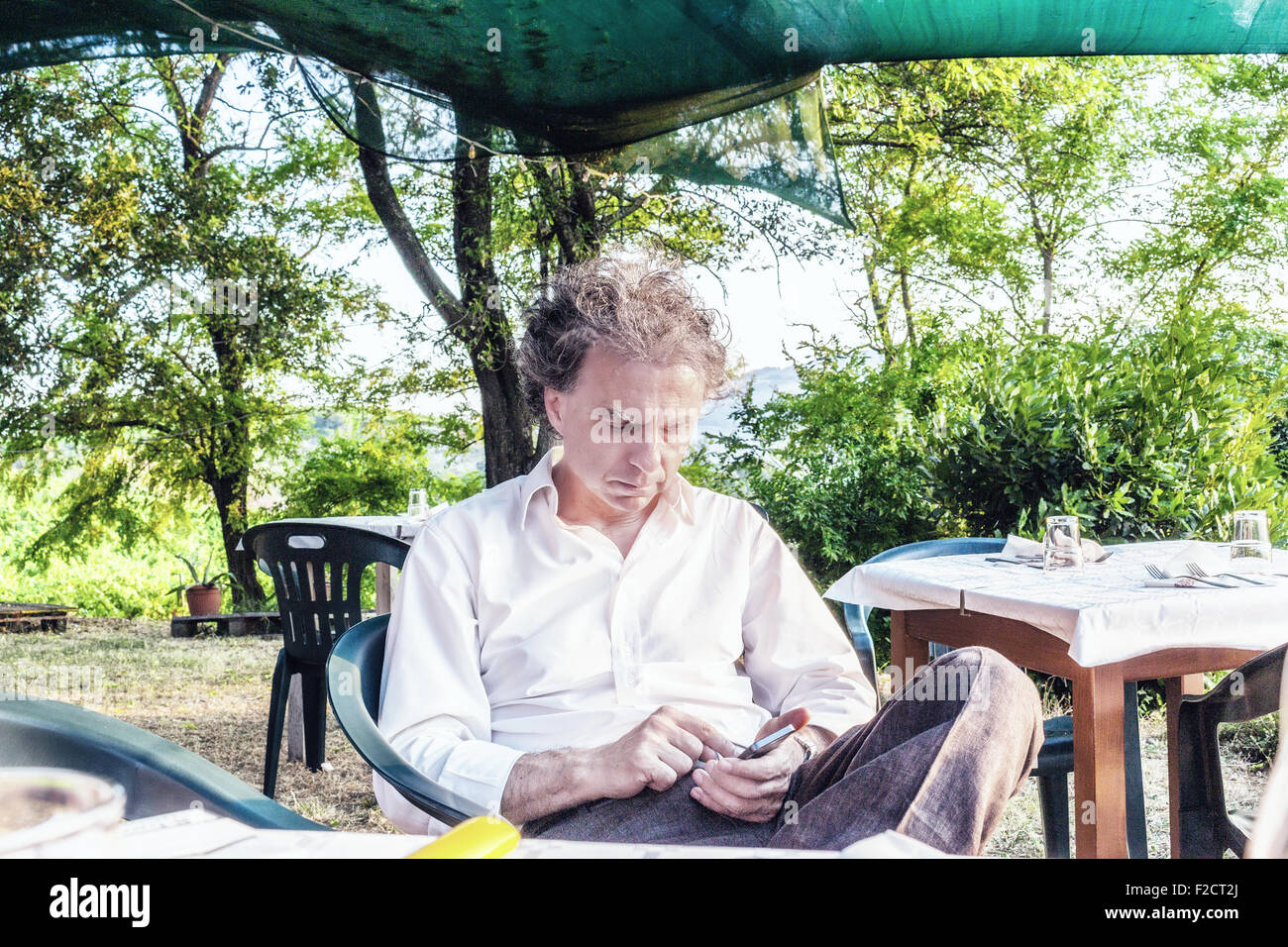 faszinierende wohlhabenden Gentleman von vierzig aus Stadt ruht auf einem Stuhl im Garten von einem roten Landhaus in Italien während mit smartphone Stockfoto