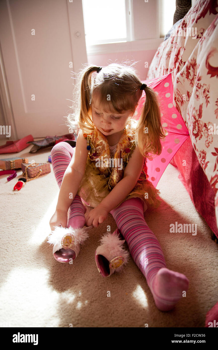 Junges Mädchen in rosa spielt setzt sich auf Boden mit hellem Licht hinter ihr und versucht auf Schuh, spielen verkleiden in ihrem Schlafzimmer Stockfoto