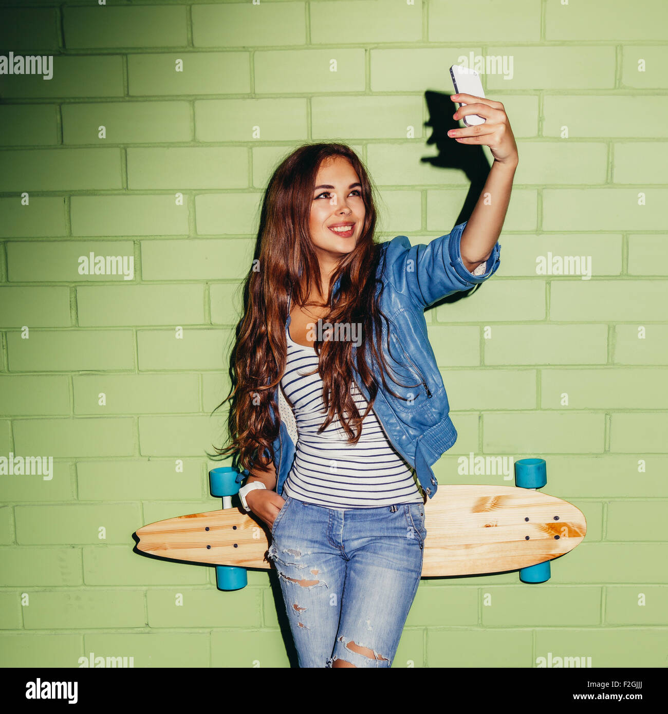 junge schöne langhaarige Brünette Mädchen mit hölzernen Longboard Skateboard machen Selfie Selbstportrait auf ihrem Smartphone digital Stockfoto