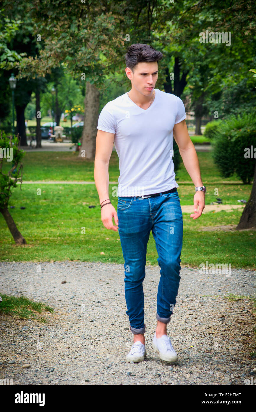 Hübscher junger Mann im weißen T-shirt und Jeans im Stadtpark, im Freien zu Fuß in Richtung Kamera, ernst Stockfoto