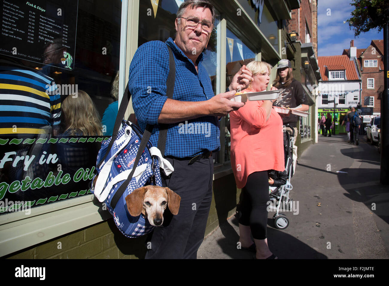 Mann isst sein Mittagessen außerhalb ein Fish &amp; Chips-Shop, mit seinem Dackel "Wurst" in seiner Tasche. Whitby, Yorkshire, Großbritannien. Stockfoto