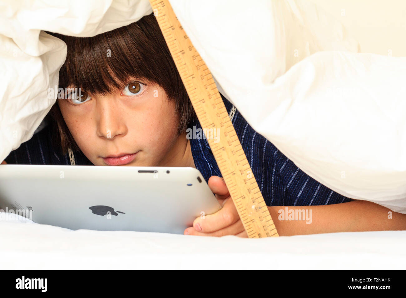 Kaukasier Kind, Junge, 10 - 11 Jahre alt. Die Hälfte unter weißen Bettdecke verstecken abgestützt durch ein Lineal. Kopf und Schultern des Jungen, und er verwendet auch das ipad, Augenkontakt. Stockfoto