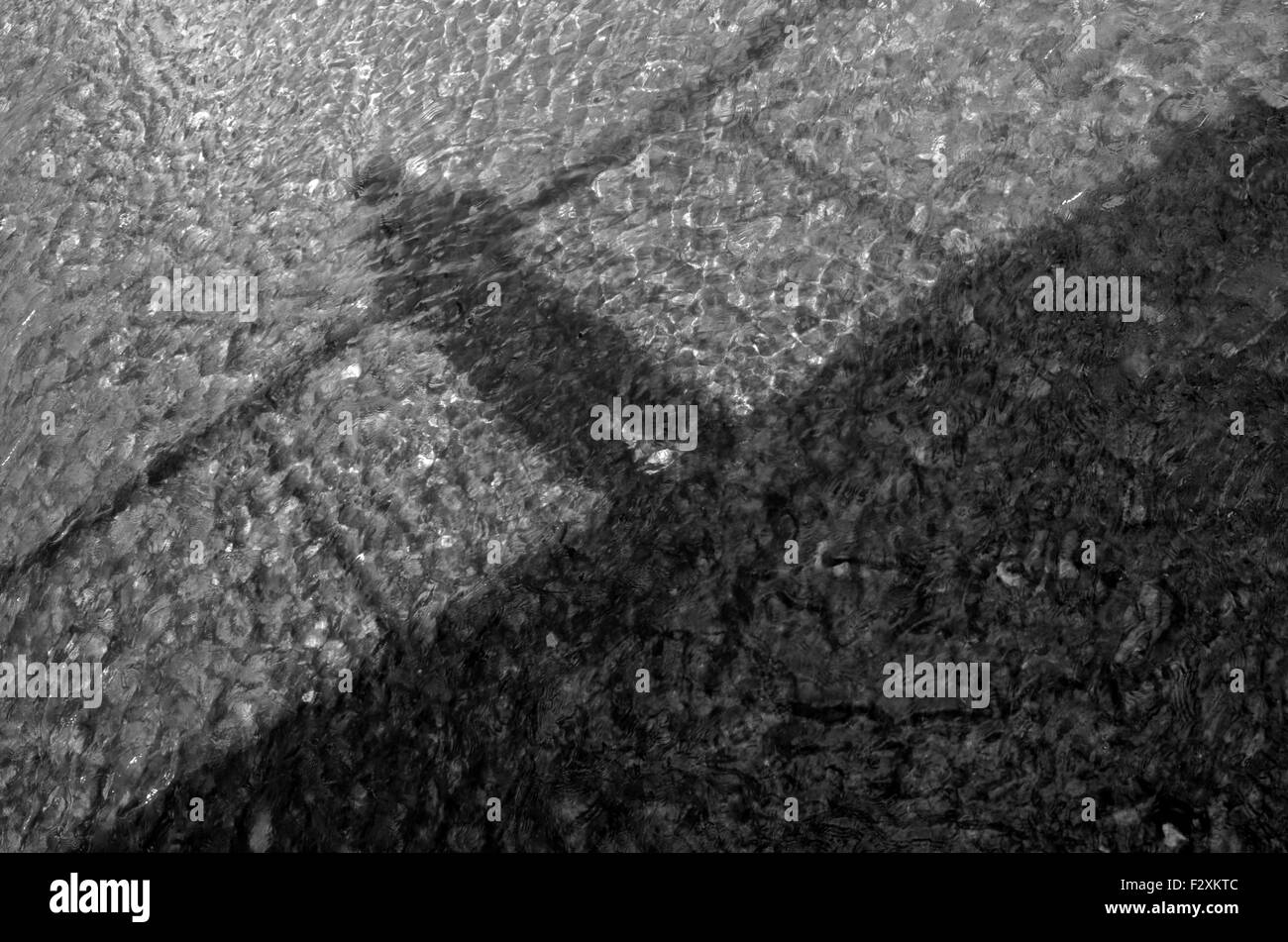 Ein monochromes Bild des Schattens eines Mannes auf einer Brücke in einen seichten Fluss wider. Stockfoto