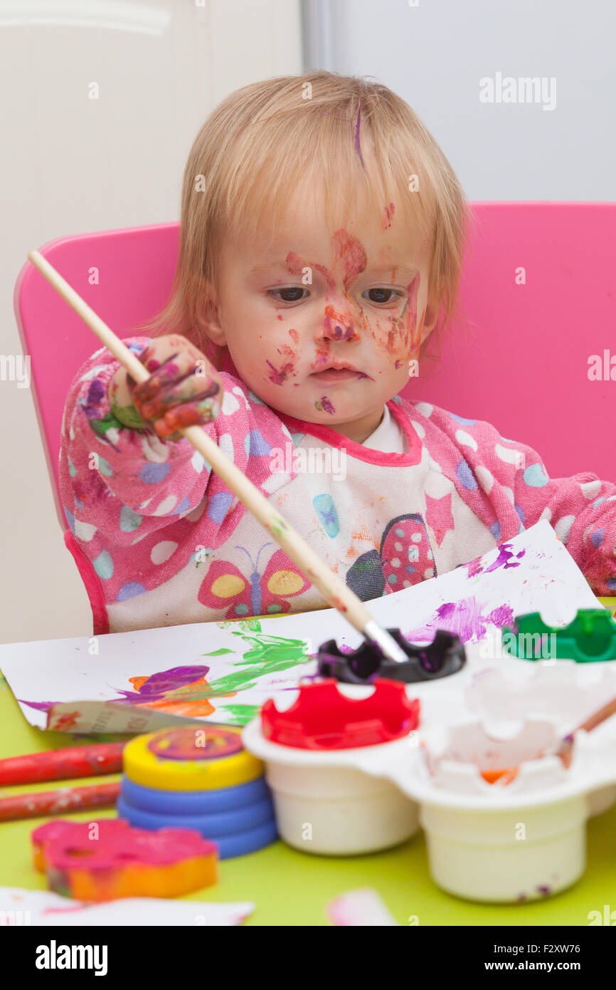 Ein 16 Monate alt, Blond Haare, Bürsten kaukasisch, Mädchen spielen mit Farben und die Farbe während des Sitzens im Inneren an einem kleinen Tisch. Stockfoto
