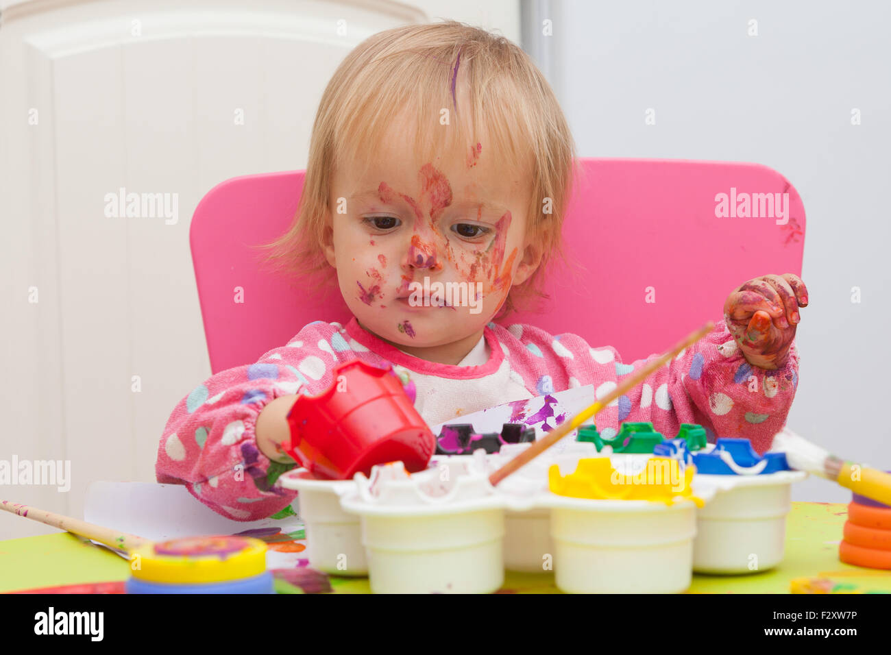 Ein 16 Monate alt, Blond Haare, Bürsten kaukasisch, Mädchen spielen mit Farben und die Farbe während des Sitzens im Inneren an einem kleinen Tisch. Stockfoto