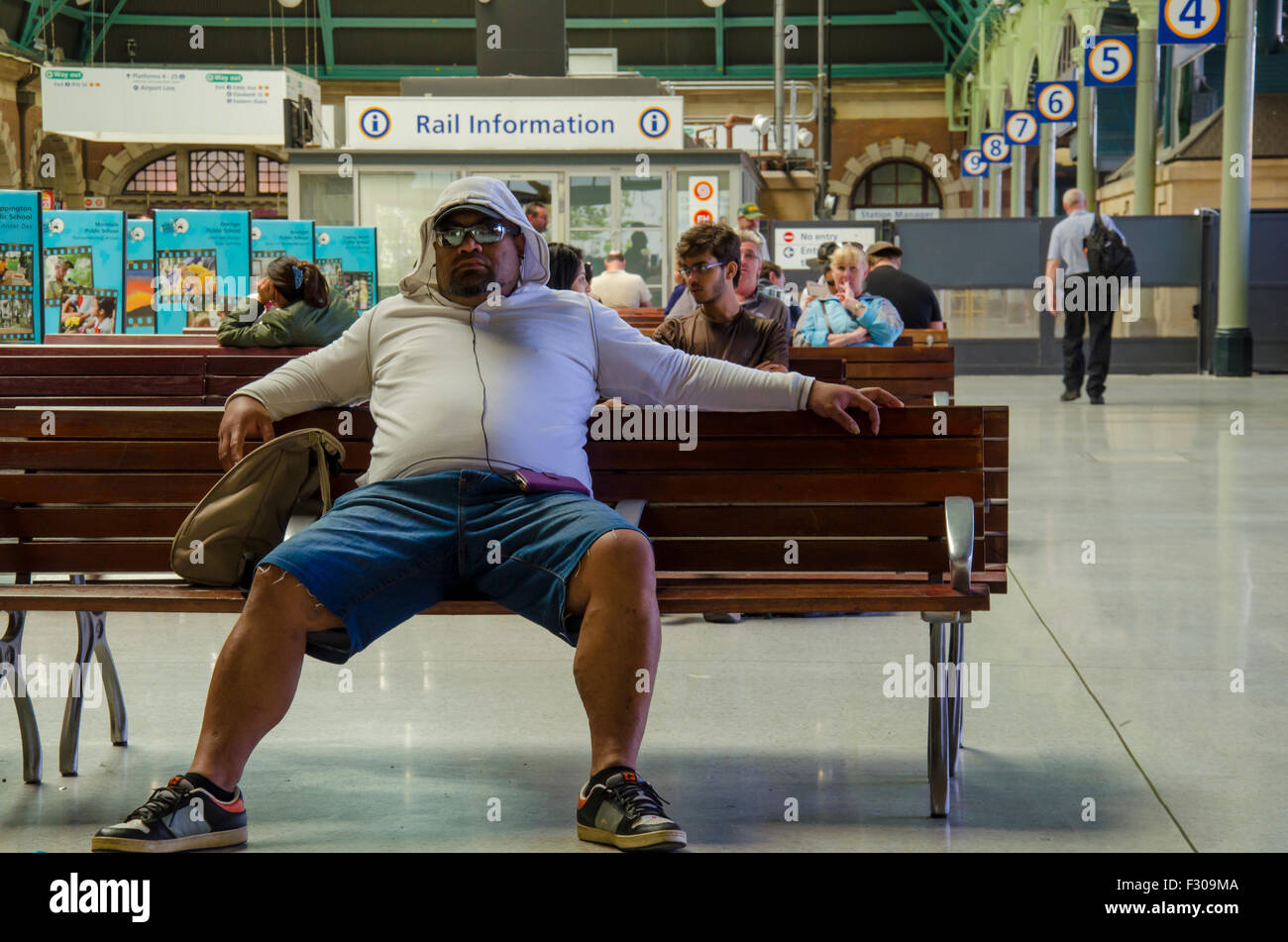 Ein übergewichtiger Mann sitzt auf einer Sitzbank mit gespreizten Beinen auf dem Landeisenbahnabschnitt des Hauptbahnhofs von Sydney in New South Wales, Australien Stockfoto