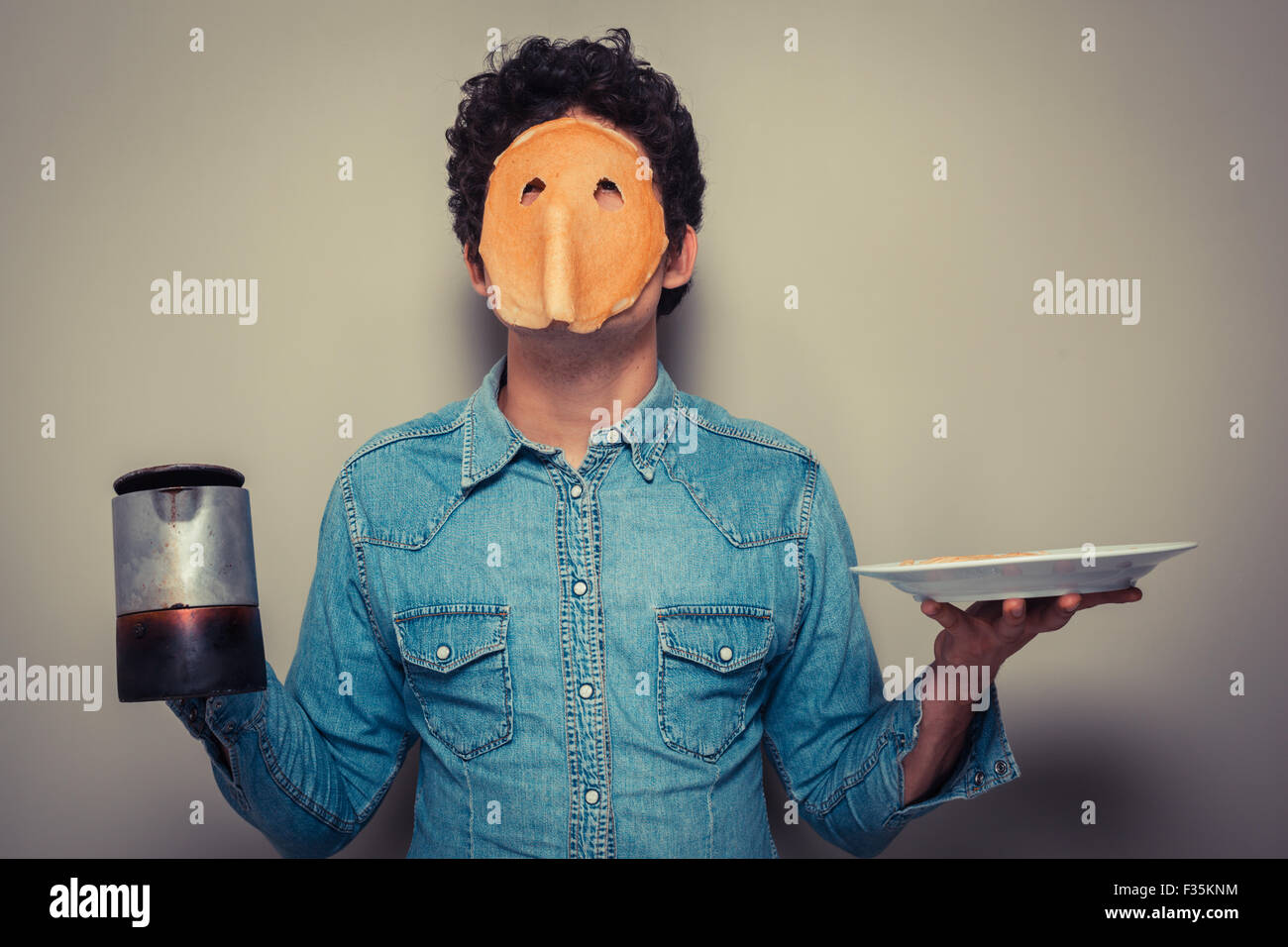 Junger Mann hat gebt in einen Pfannkuchen geschnitten und trägt es auf seinem Gesicht halten Sie eine Kanne Kaffee Stockfoto