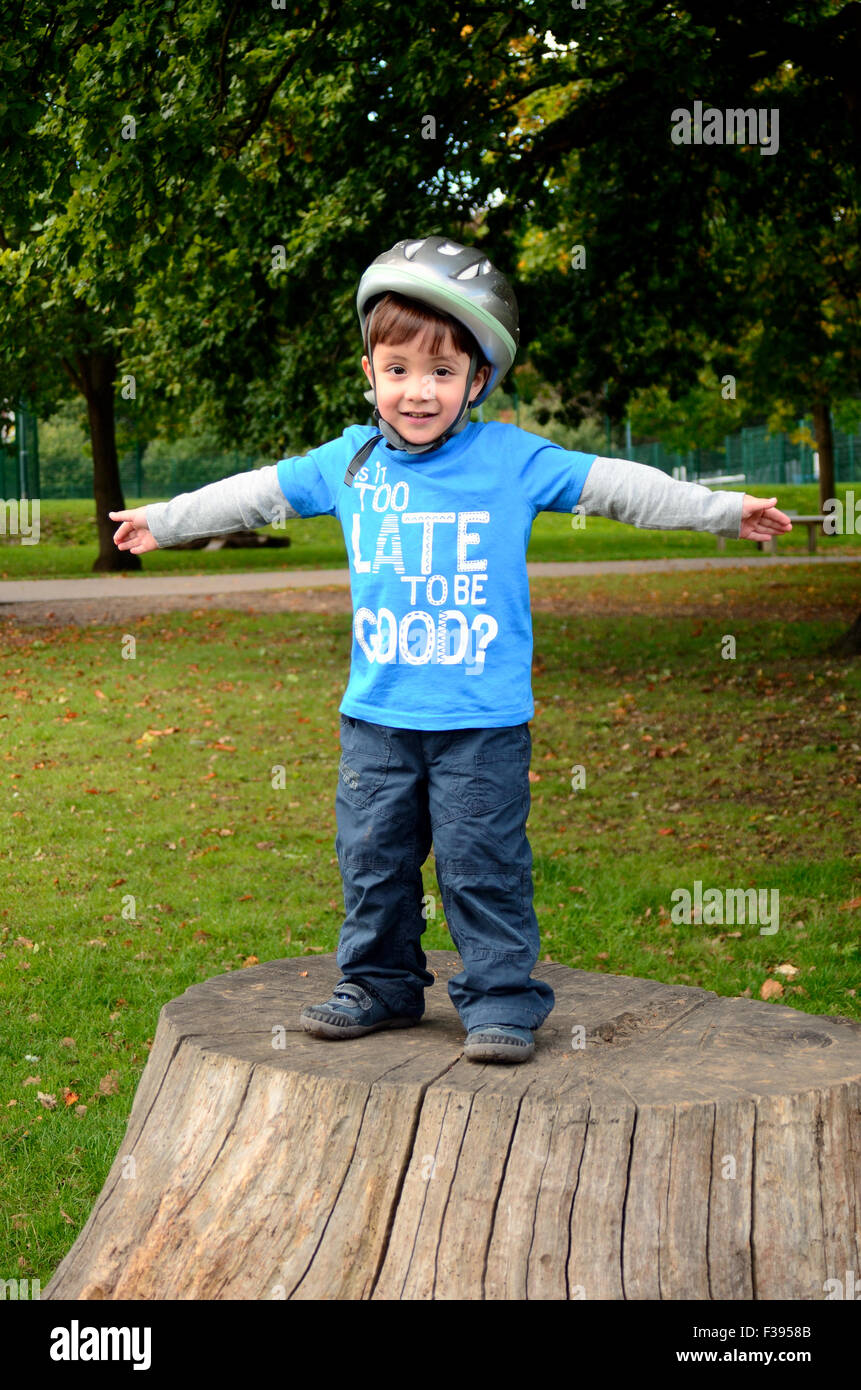 Ein kleiner Junge, das Tragen eines Helms beim Radfahren steht auf einem Baumstumpf im Park. Stockfoto