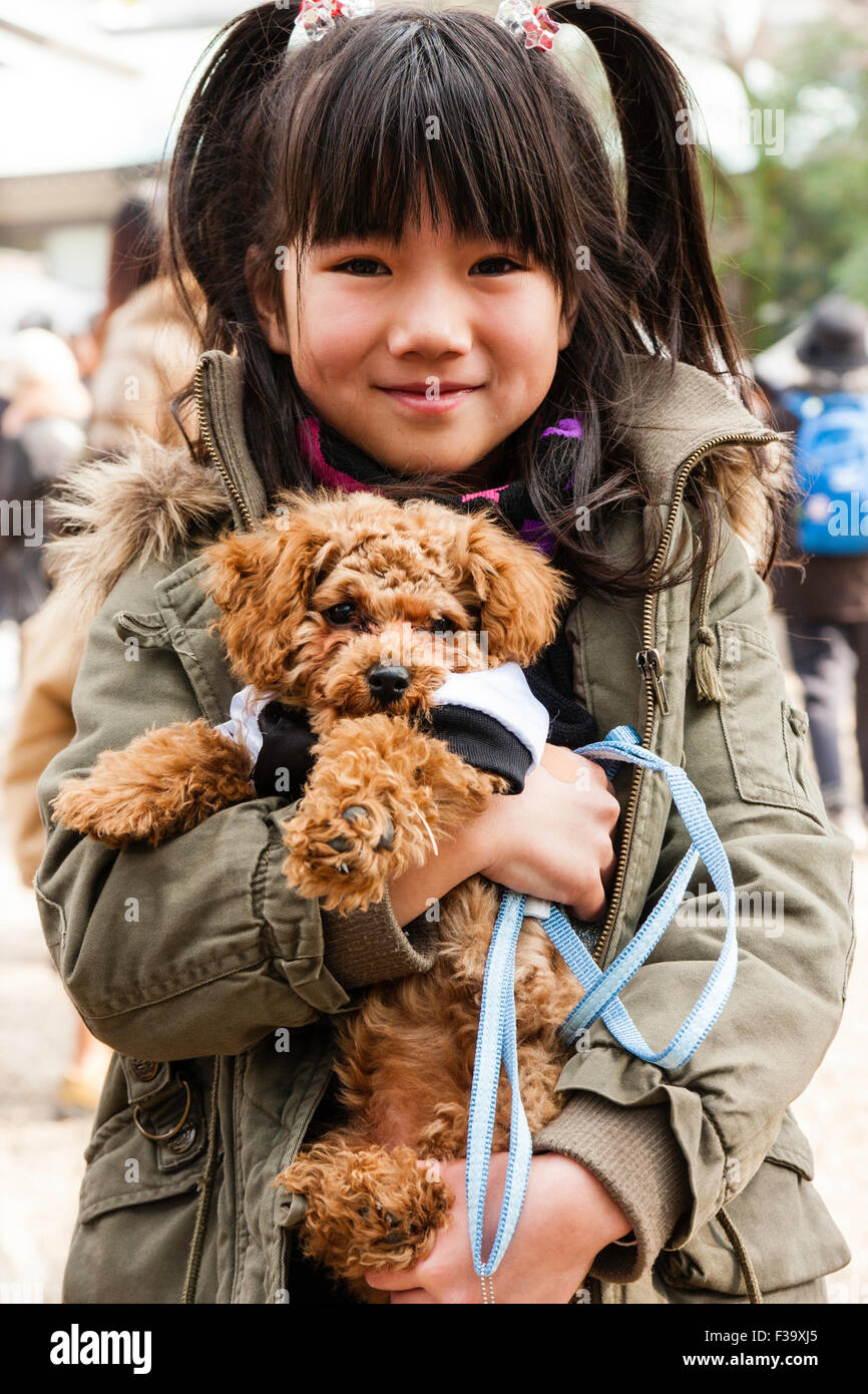 Japanisches Kind, Mädchen, 9-10 Jahre alt, in Mantel, lächeln und posieren für Viewer während sie Streicheleinheiten und hält in beiden Armen ihr Haustier Hund. Auge - Kontakt. Stockfoto