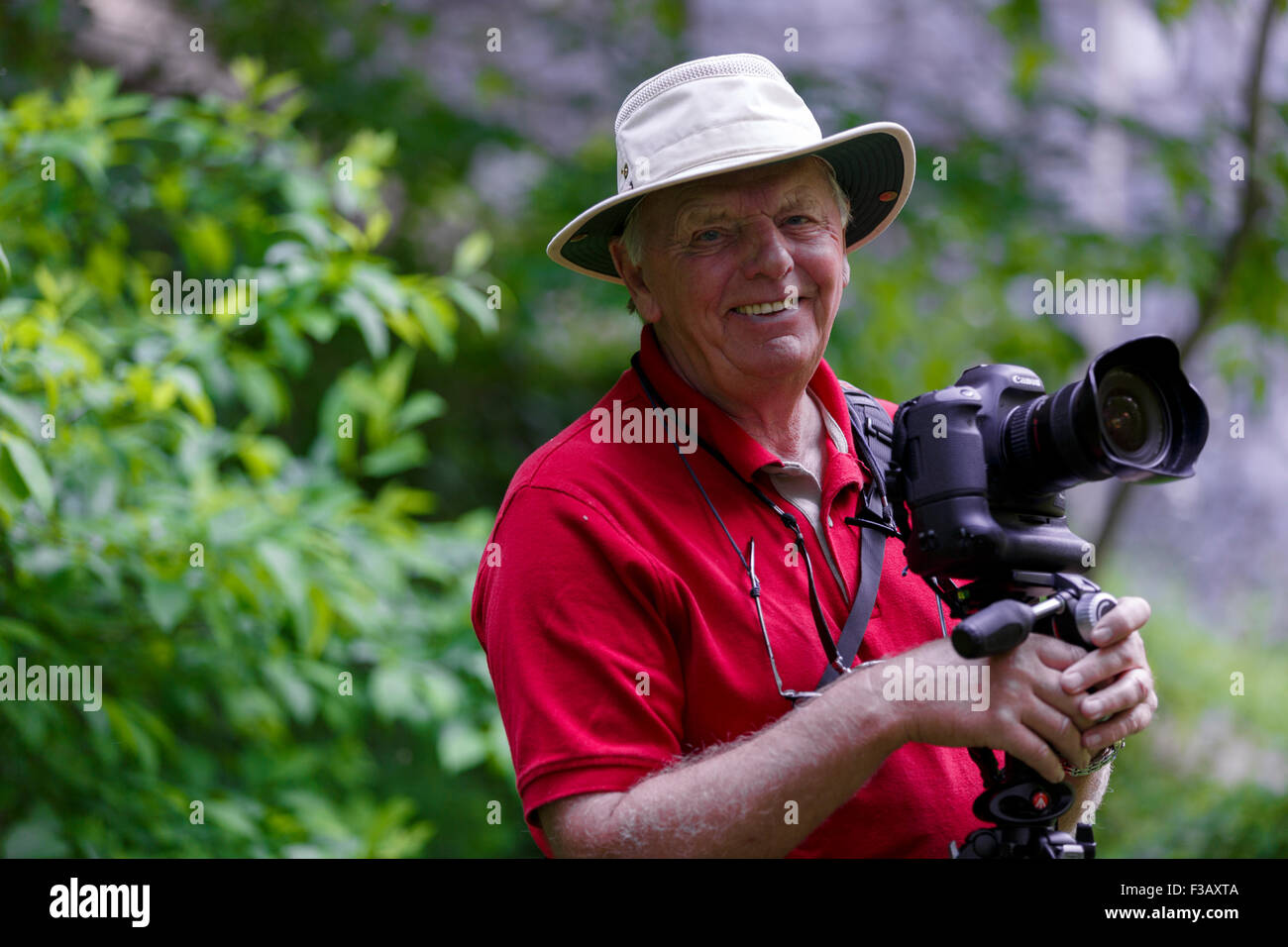 Lächelnder älterer Mann, der Tilley hat trägt und die Kamera lächelnd anschaut Fotograf verschwommen Laub Hintergrund Stockfoto