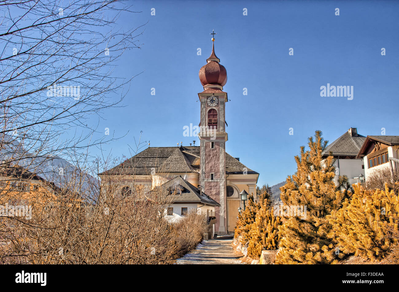 Der Luis Trenker Promenade führt bis St. Ulrich, italienisches Dorf in Dolomiten Alpen: typische Häuser, Gebäude und die roten Glockenturm mit Uhr Pfarrkirche St. Ulrich mit Bergen und grünen Koniferen im Hintergrund Stockfoto