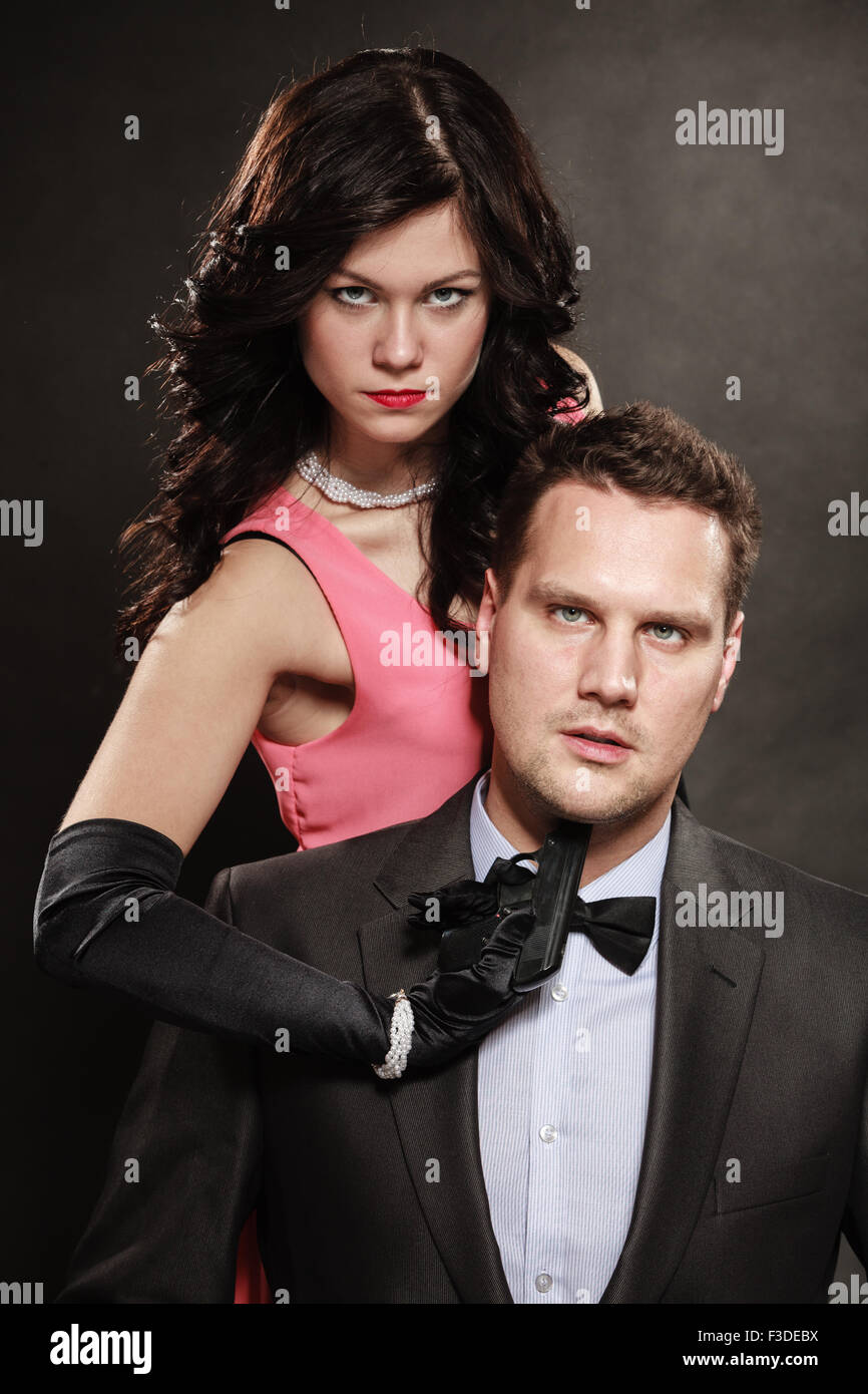 Szene der Gewalt mit Schusswaffe zwischen Männern und Frauen. Elegante Dame mit Pistole gegen sitzen Mann im Anzug auf schwarz und grau bac Stockfoto