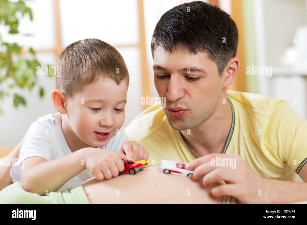 Fröhliche Vater und Sohn haben ein lustiges Spiel mit Spielzeugauto auf Bauch der schwangeren Frau. Stockfoto