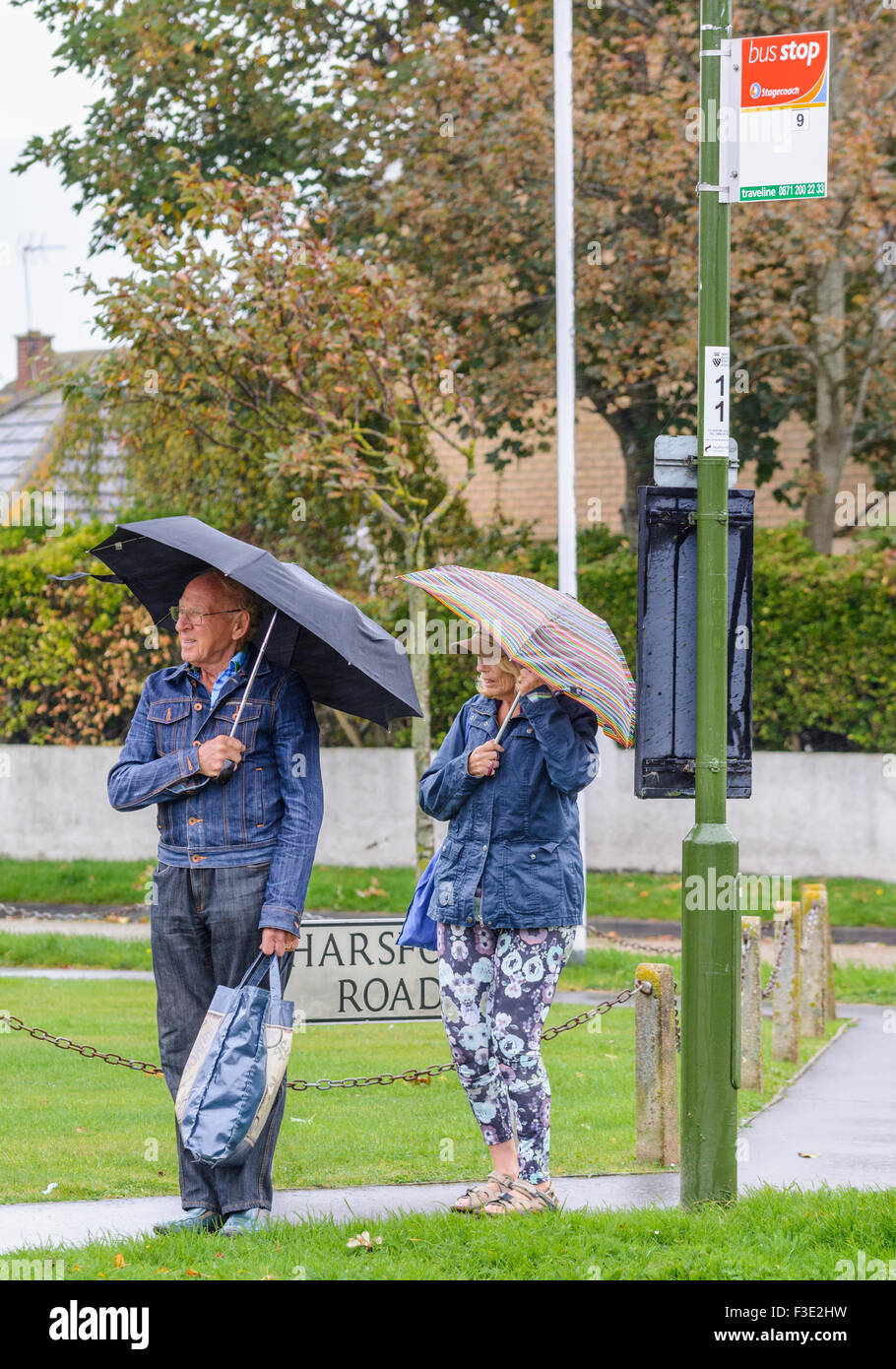 Ältere Paare warten an einer Bushaltestelle holding Regenschirme im Regen, in Großbritannien. Stockfoto