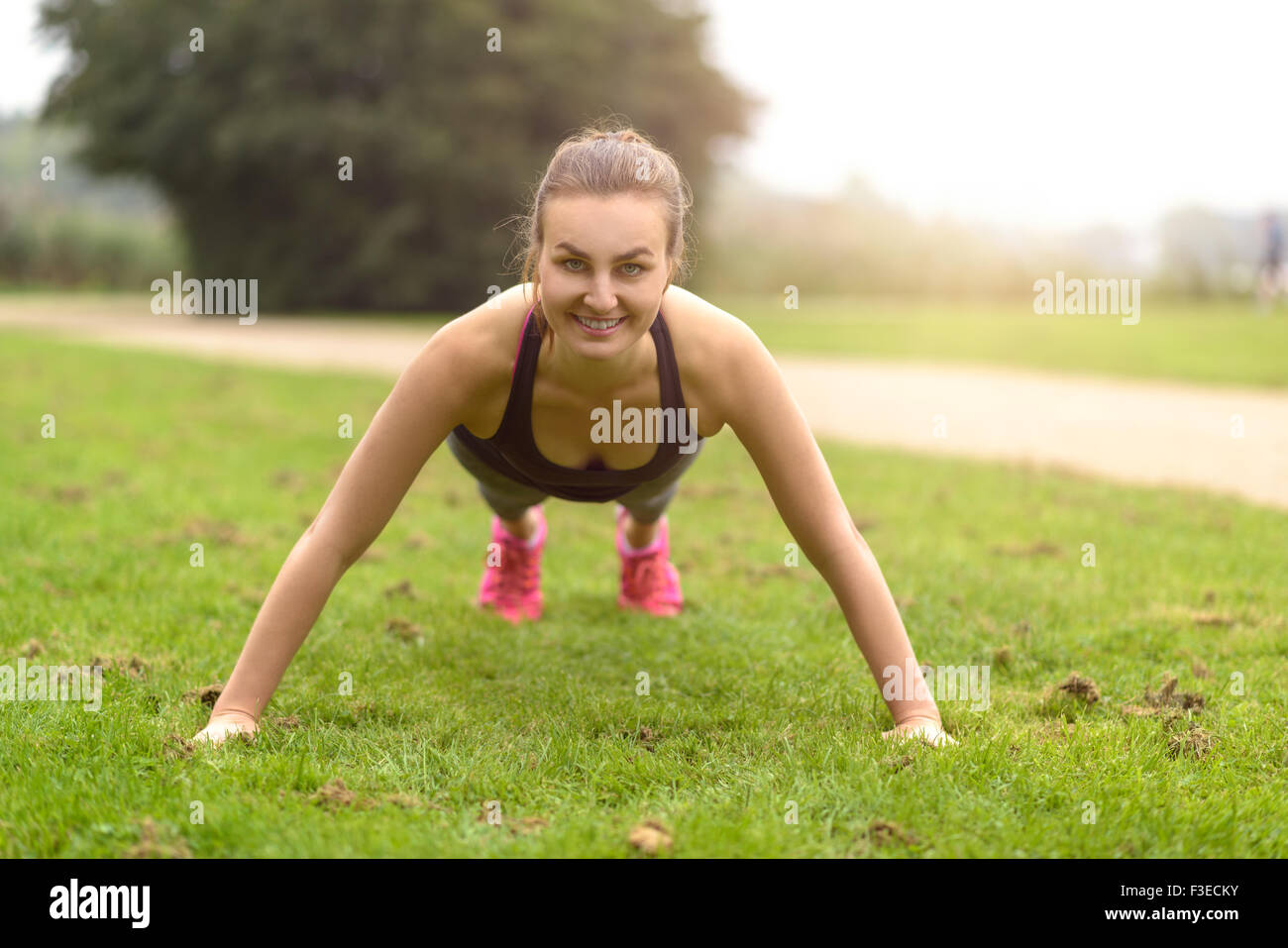 Sportliche junge Frau lächelt in die Kamera dabei Push-Up-Übung im Park. Stockfoto