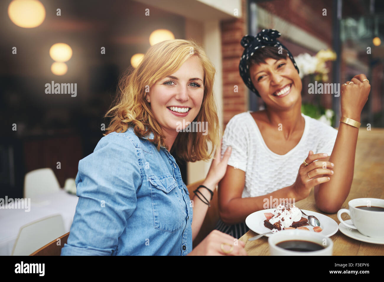 Zwei attraktive junge multiethnischen Freundinnen genießen Sie Kaffee und Kuchen zusammen in einer Cafeteria glücklich lächelnd in die Kamera ein Stockfoto