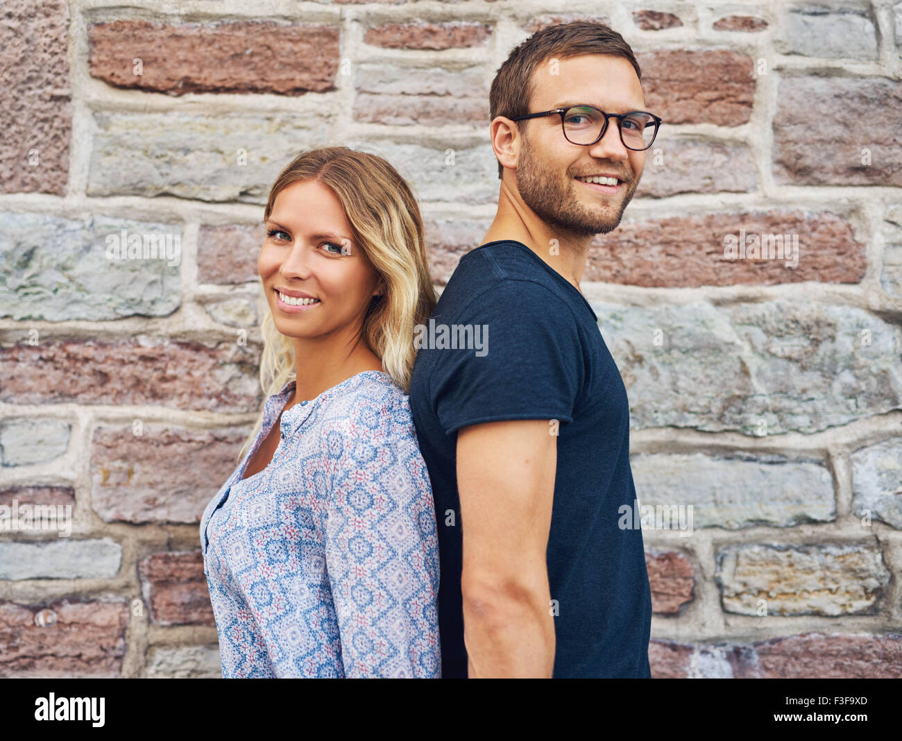 Glückliches Paar stehen Rücken an Rücken und lächelnd in die Kamera vor Brick Wall Hintergrund Stockfoto