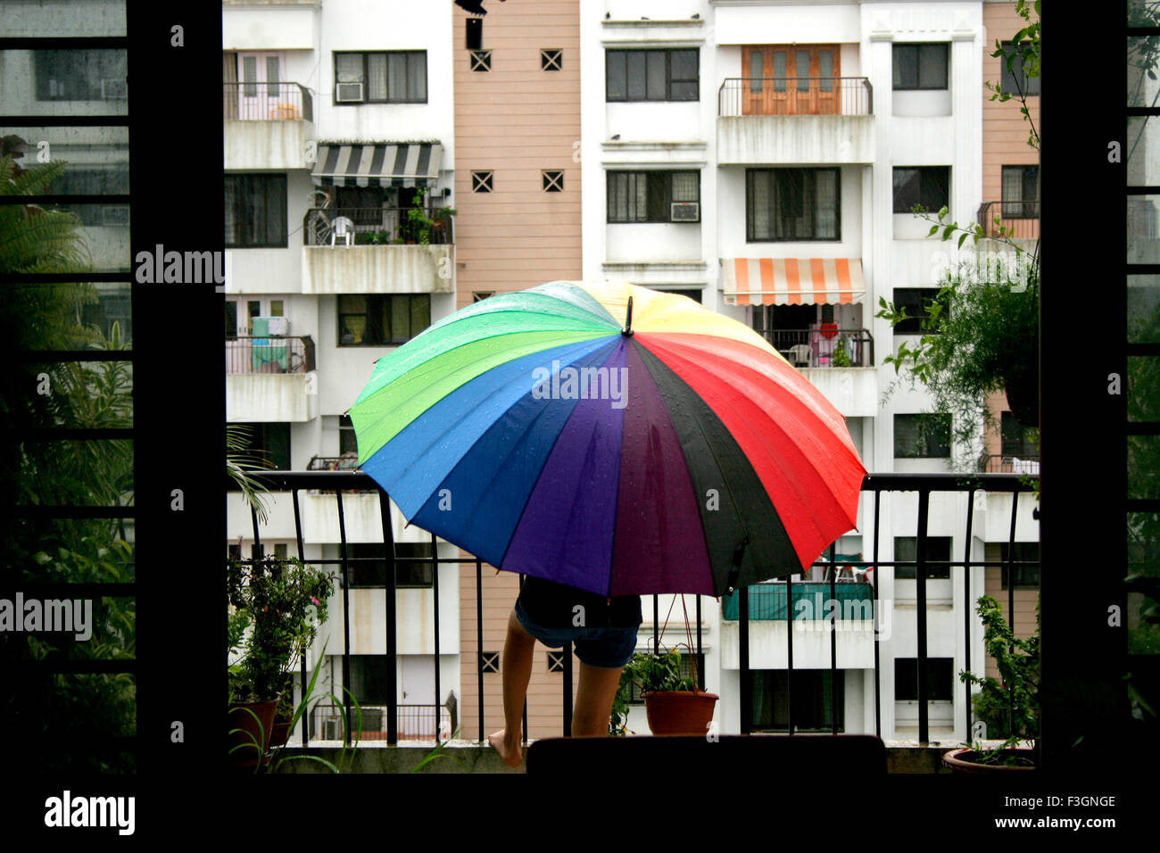 Ein indisches Mädchen Kind spielt auf dem Balkon ihres Hauses im Monsun mit einen großen sechzehn farbigen weit geöffneten Regenschirm Stockfoto