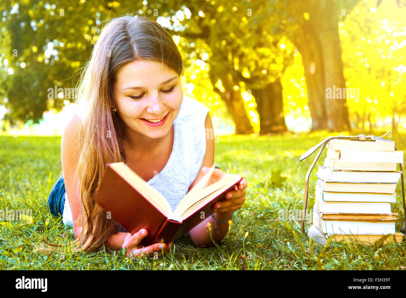 Junge schöne College-Studentin, liegend auf dem grünen Rasen und einem Buch auf dem Campus am warmen Tag. Bildung. Zurück zu Stockfoto