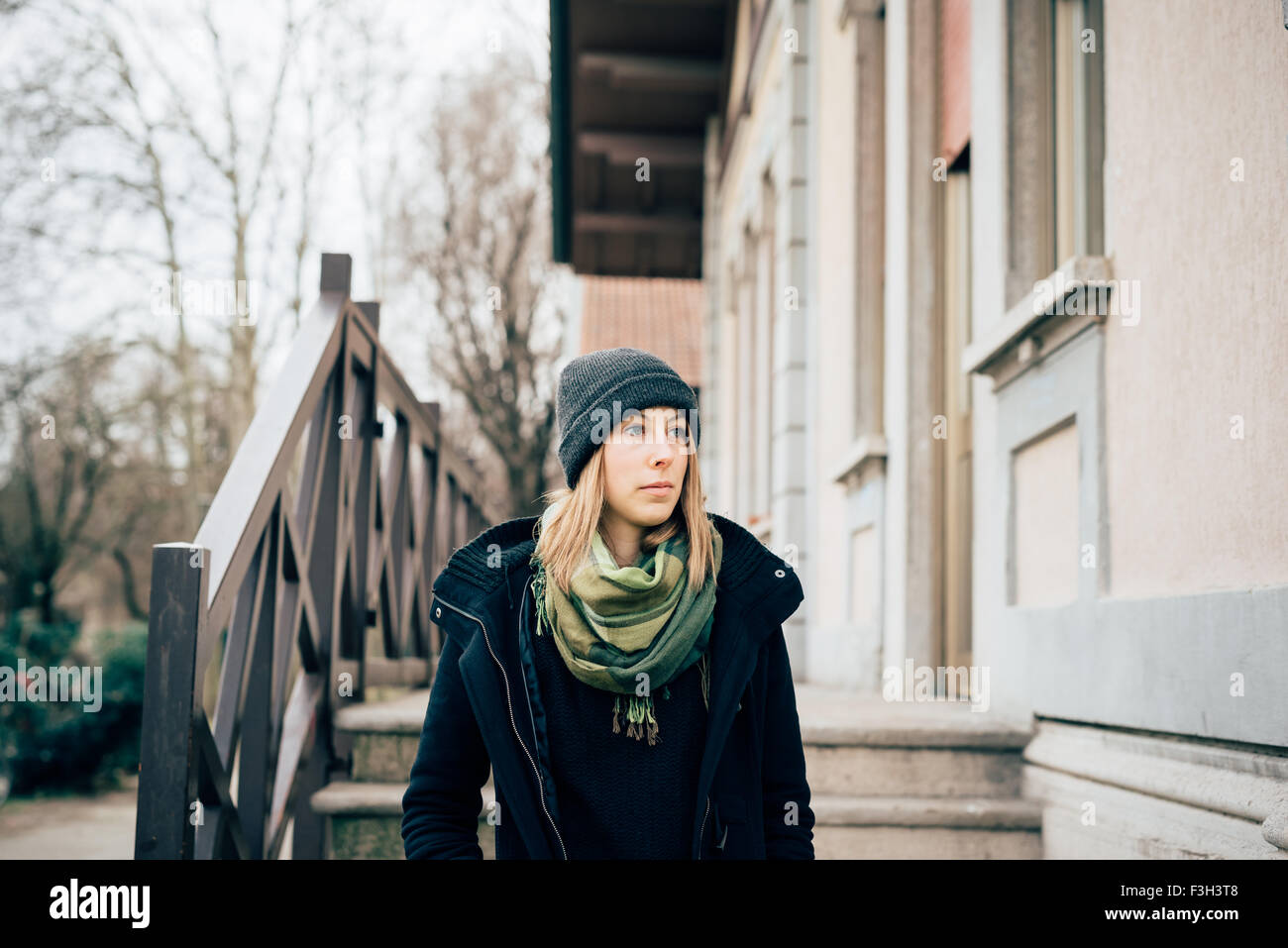 Halbe Länge einer jungen schönen Hipster sportliche blonde Frau zu Fuß in  der Stadt, die Hände in Tasche, trägt einen Hut, einen grünen Schal und ein schwarzer  Pullover und Mantel. Mit Blick