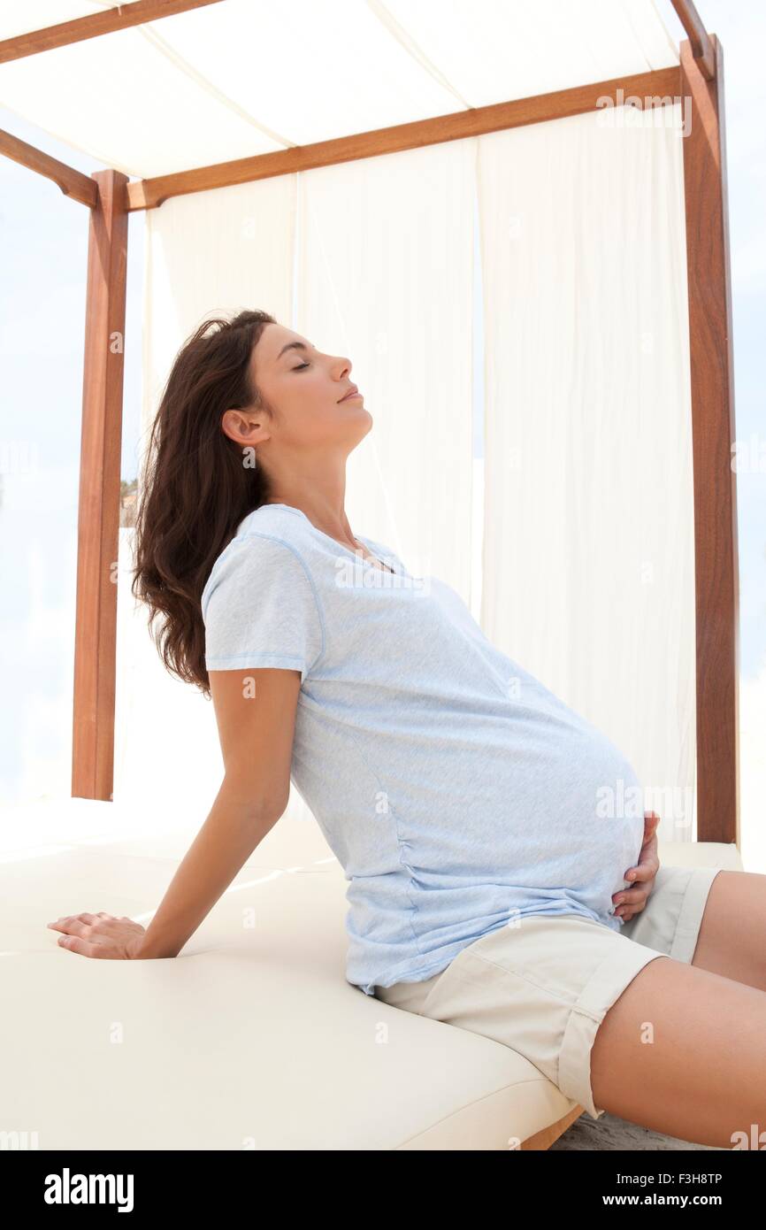 Mitte Erwachsene Frau sitzt auf der Liege mit Hand auf Bauch schwanger Stockfoto