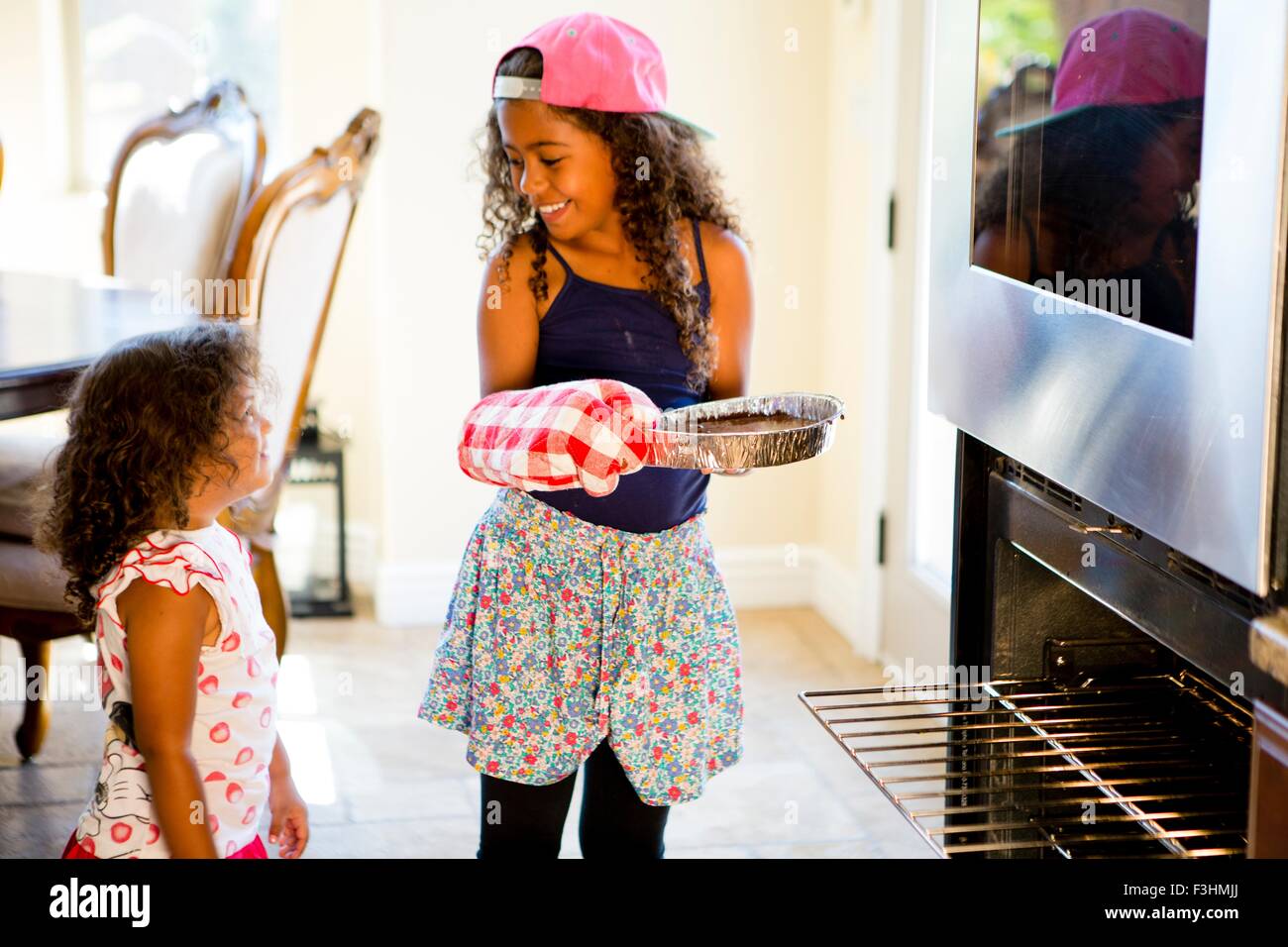 Mädchen beobachten große Schwester einfügen Kuchenform in Ofen, von Angesicht zu Angesicht lächelnd Stockfoto