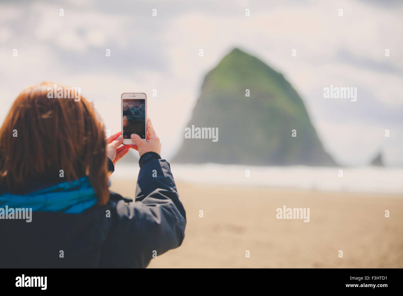 Eine junge Frau nutzt ihr Smartphone, um Haystack Rock in Cannon Beach fotografieren. Stockfoto