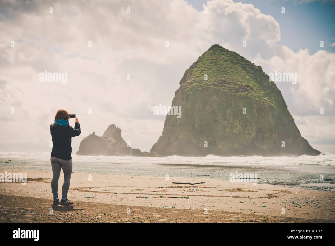 Eine junge Frau nutzt ihr Smartphone, um Haystack Rock in Cannon Beach fotografieren. Stockfoto