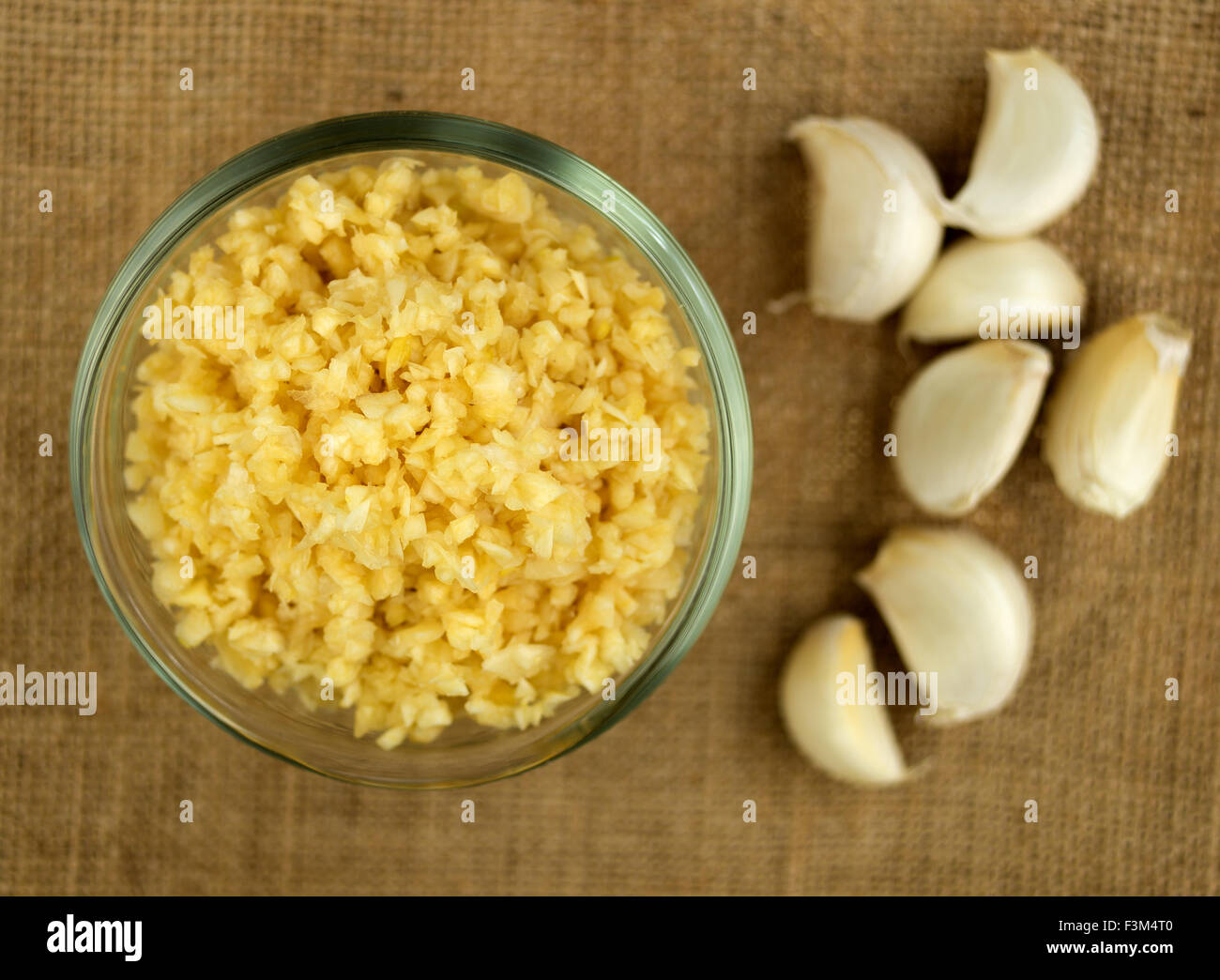 Aerial Makro fein gehackten Knoblauch in einer Schüssel gegen Gunny Tuch mit ganzen Knoblauchzehen im Hintergrund Stockfoto