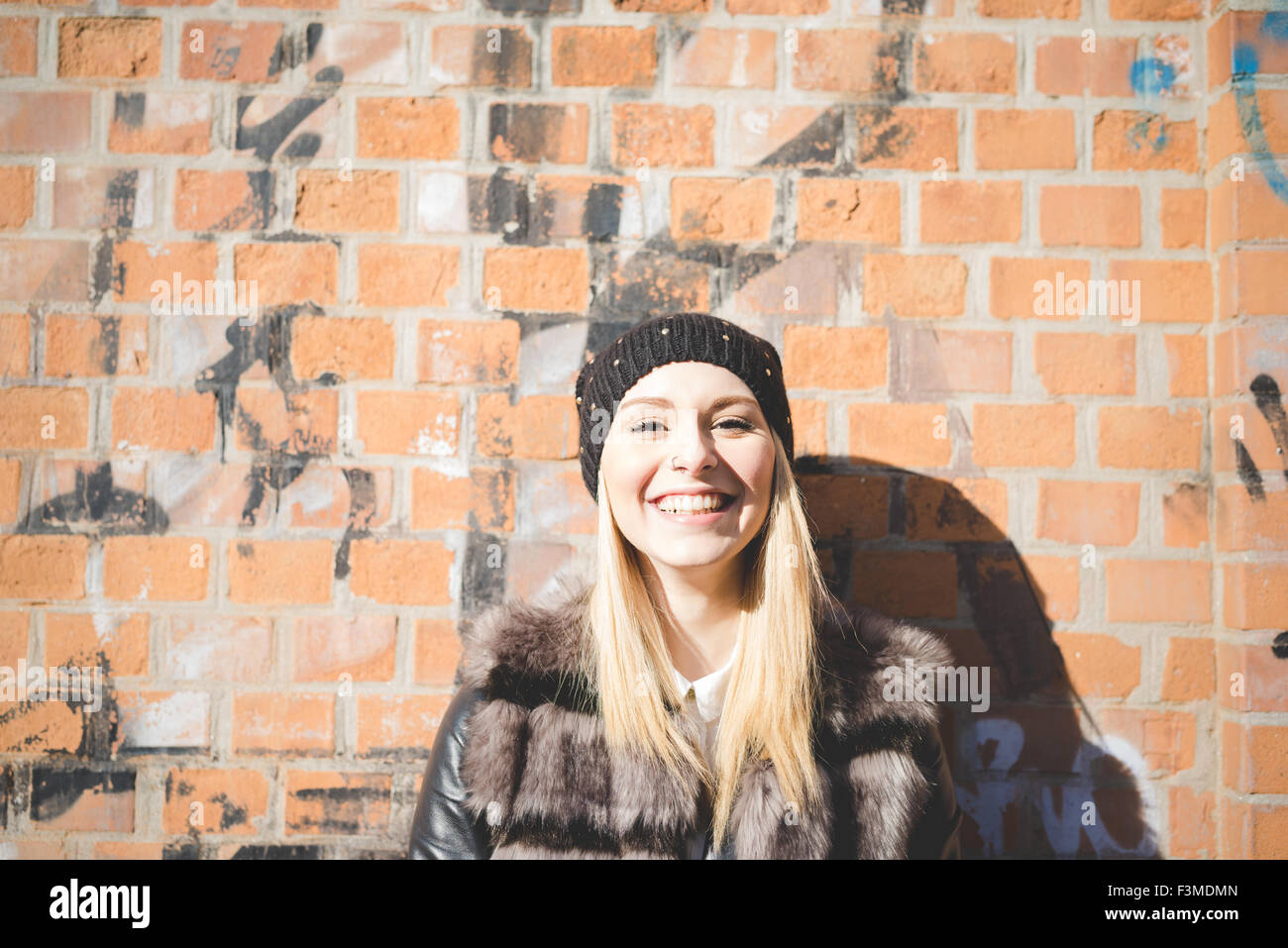 Porträt der junge schöne blonde glatte Haare Frau in der Stadt an eine Wand gelehnt in der Kamera suchen, Lachen - Glück, Sorglosigkeit, fun-Konzept - mit schwarzem Hut und pelzige braune Jacke Stockfoto