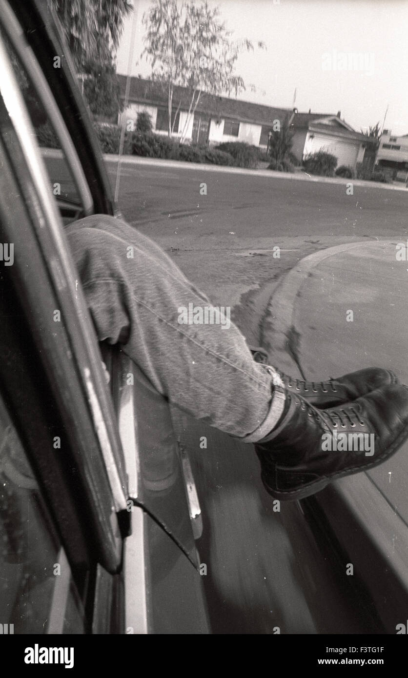 Ein punker Punk Rocker tragen Doc Marten Springerstiefel baumelt seine Beine aus einem Auto eine Ecke bog in eine s-Darm-Trakt nach Hause Nachbar Kapuze Porterville, Kalifornien der 1980er Jahre Mode Teenager junge Teenager Stockfoto