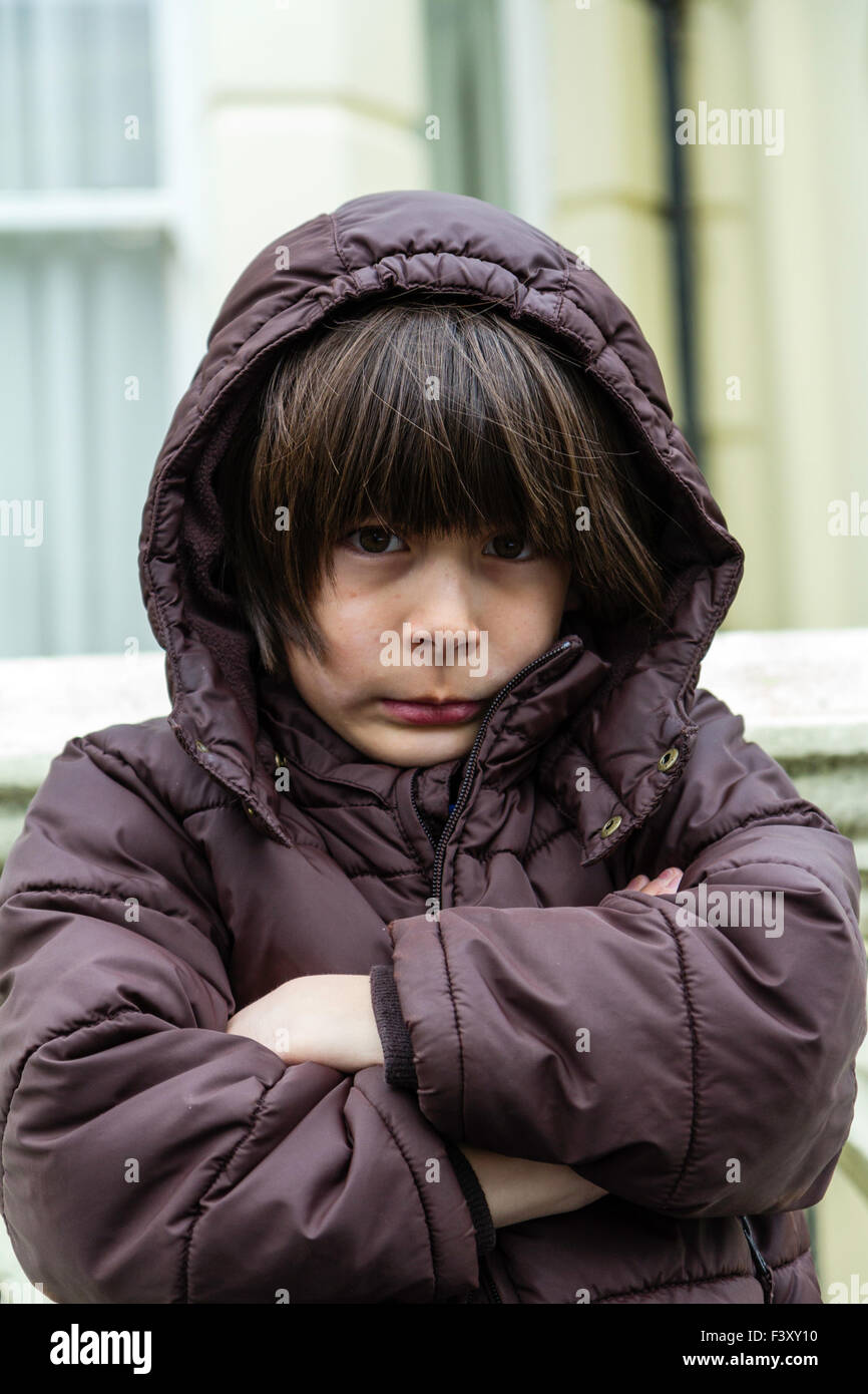 Kaukasier Kind, Junge, 9-10 Jährige, draußen stehen, tragen Anorak, kalt, die Arme verschränkt, warten, unglücklichen Ausdruck auf seinem Gesicht, Haube Stockfoto