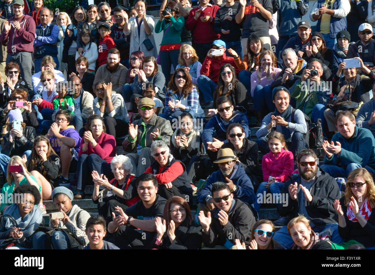 Eine große, vielfältige Gruppe multiethnischer Menschen, die draußen in Reihen sitzen, als Publikum, das Straßenkünstler beobachtet, mit fröhlichen Reaktionen in den USA Stockfoto