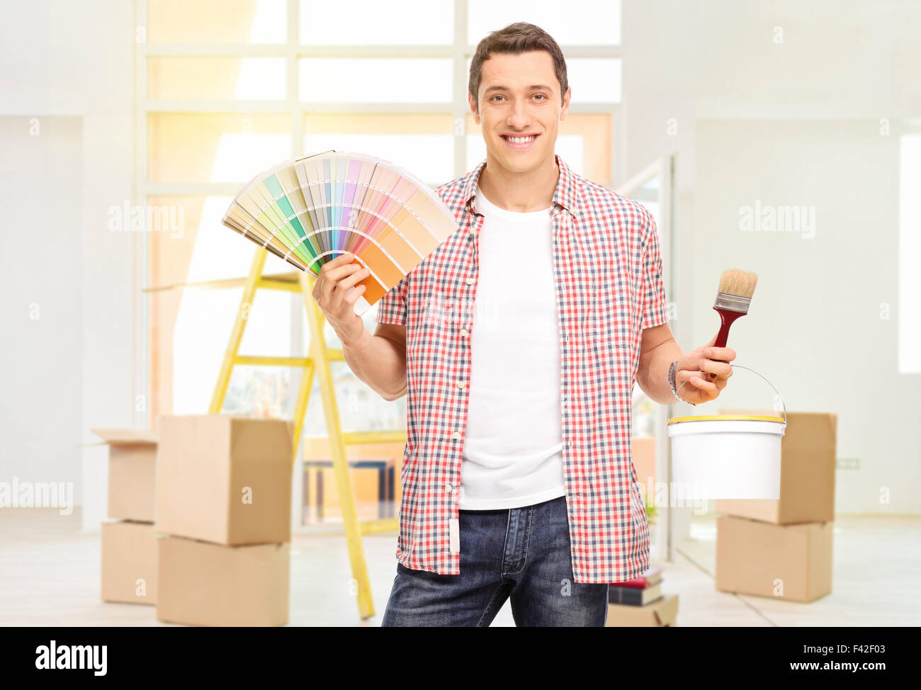 Fröhlicher junger Mann hält einen Pinsel und ein Farbfeld in einer Wohnung Stockfoto