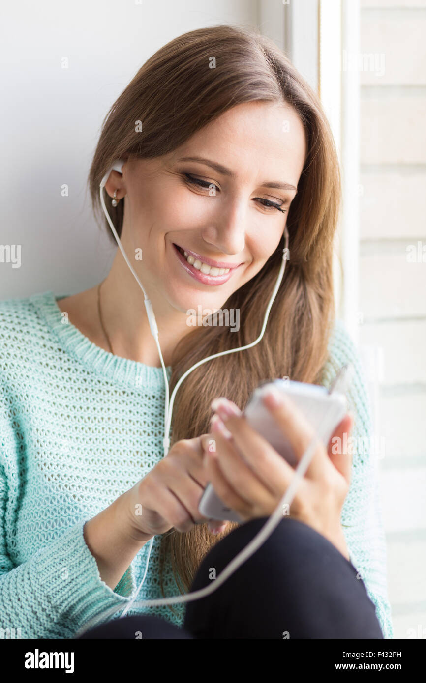 Junge lächelnde Frau mit Spaß Musik hören und lernen auf ihrem Smartphone. Glücklich schöne Mädchen am Fenster Si entspannend Stockfoto