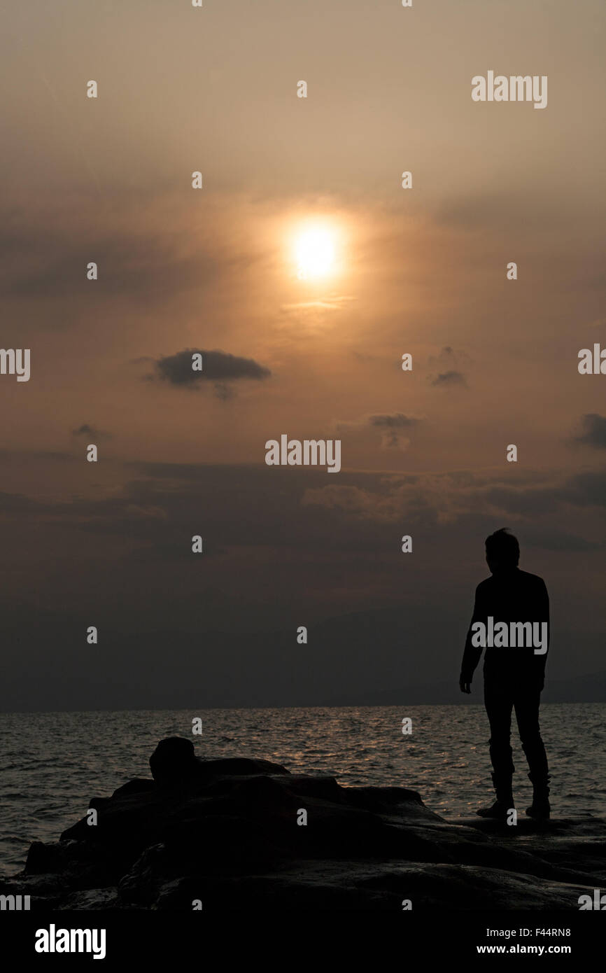 Silhouette des Menschen auf Felsen über dem Meer die Sonne niedrig am Himmel hinter Wolke Dunst Stockfoto