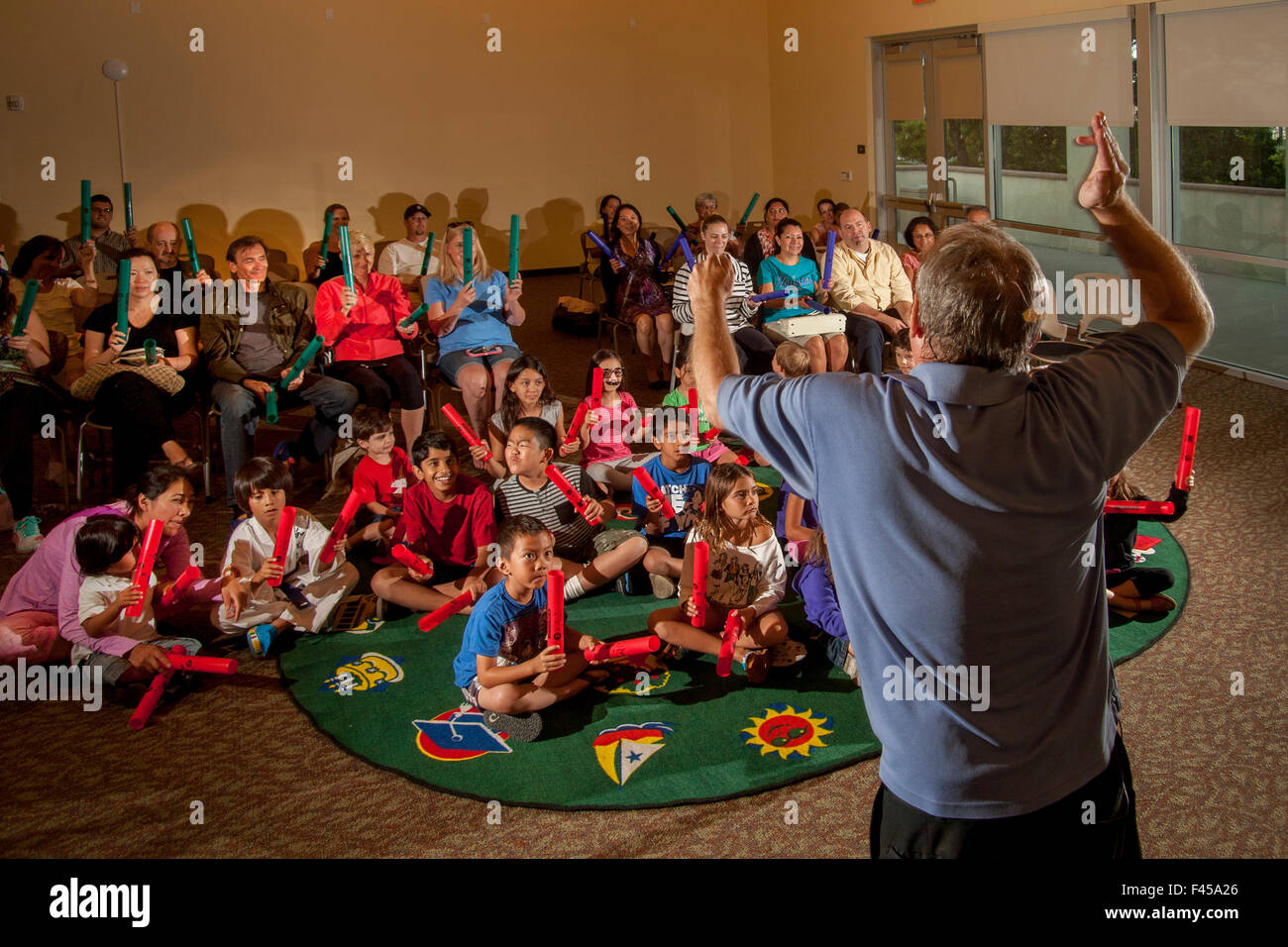 Eine multirassische Kinder Rhythm Band spielt tuned Percussion Röhren Boomwhackers am Laguna Niguel, CA, Stadtbibliothek Mehrzweckraum aufgerufen. Beachten Sie die Eltern im Hintergrund. Hinweis Erwachsenen Marktführer im Vordergrund Stockfoto
