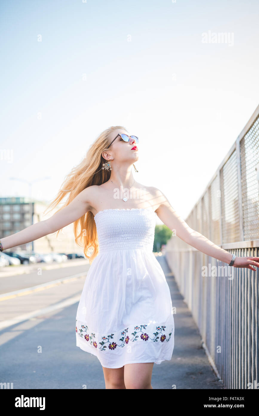 Knie Figur des jungen schönen kaukasischen langes blondes glattes Haar Frau zu Fuß in die Stadt, mit Blick auf Links, mit weit geöffneten Armen frei fühlen - weißen Kleid - Freiheit und Sorglosigkeit Konzept Stockfoto