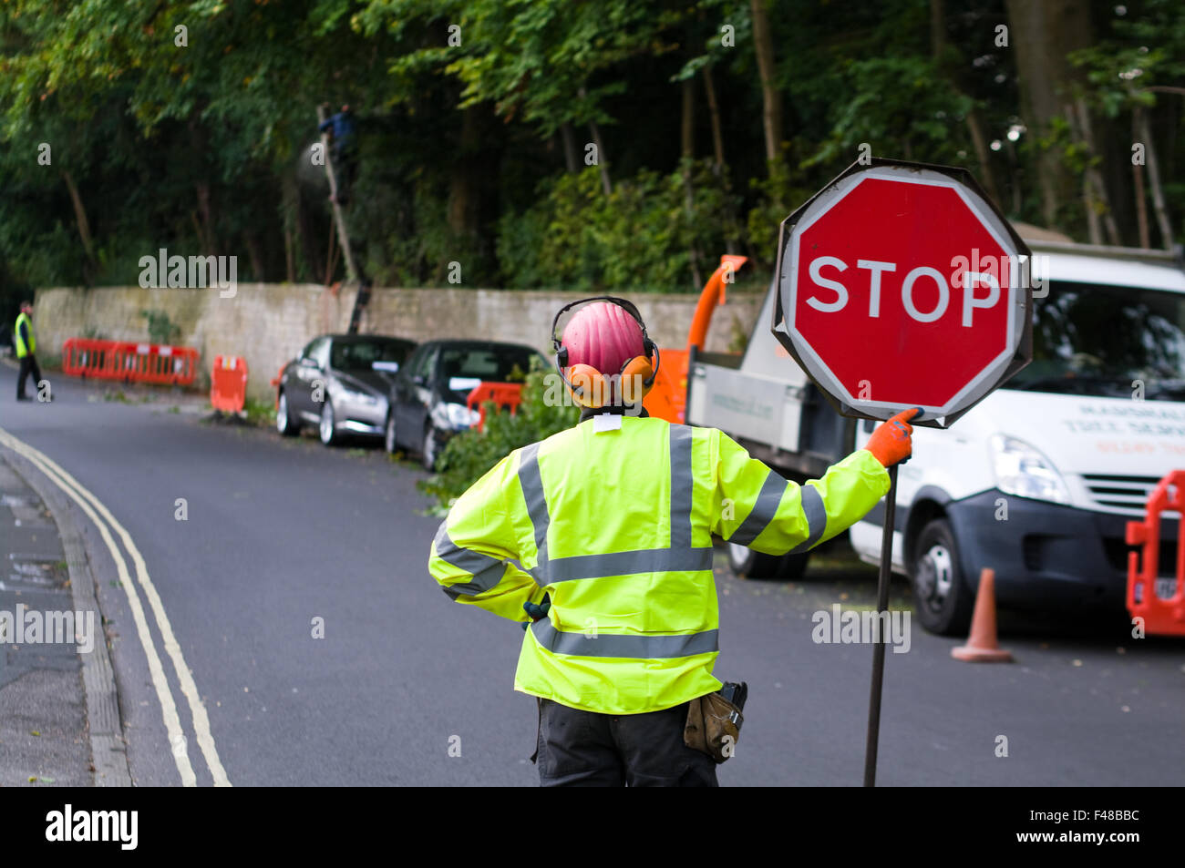 Ein Mann in eine hohe Sichtbarkeit Jacke hält ein "STOP" Kontrolle Verkehrszeichen, Verkehr zu kontrollieren, wie Mitarbeiter auf Baumschlagen arbeiten. Stockfoto