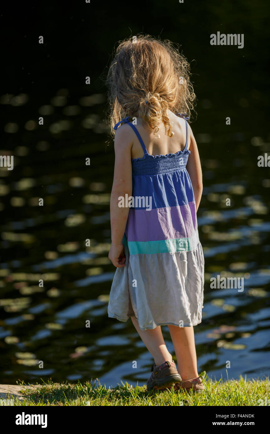 Junges Mädchen im Sommer Kleid mit offenen langen Haaren steht mit Rücken zur Kamera auf dem Rasen am Gewässerrand Stockfoto