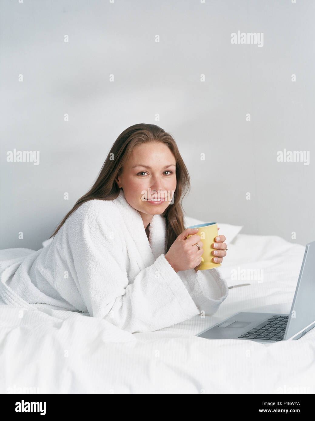 30-34 Jahre Erwachsene nur Bademantel Bett Farbe Bild Computer verbunden Tasse trinken Essen und trinken freundlich Vorderansicht glücklich im Innenbereich Stockfoto